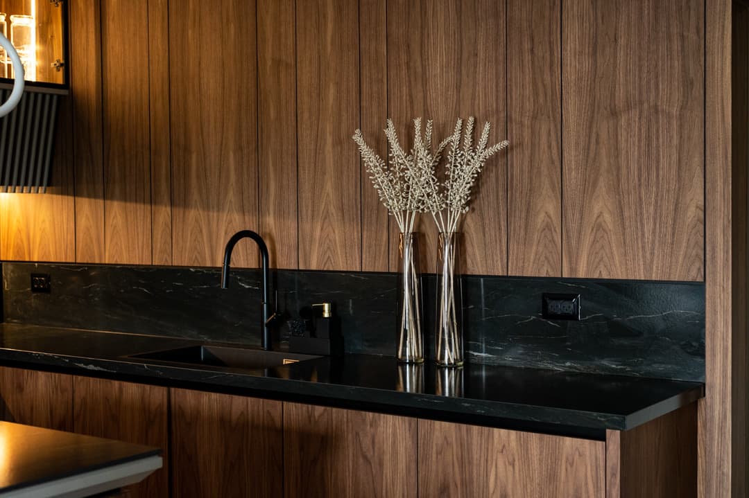 Black gooseneck tap and dried grass vases on a black stone counter against walnut panelling in the kitchen of Marquina