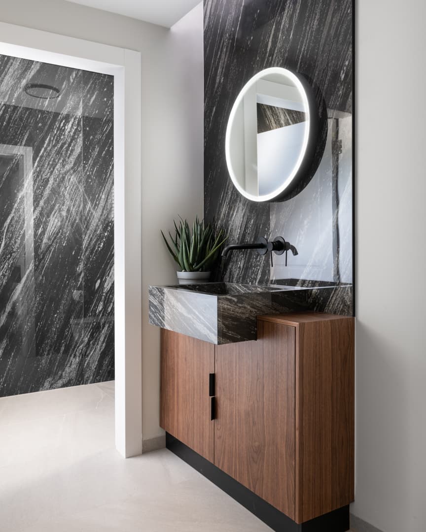 Black streaked stone sink on a walnut cabinet with a backlit round mirror and black wall tap in the powder room of Marquina