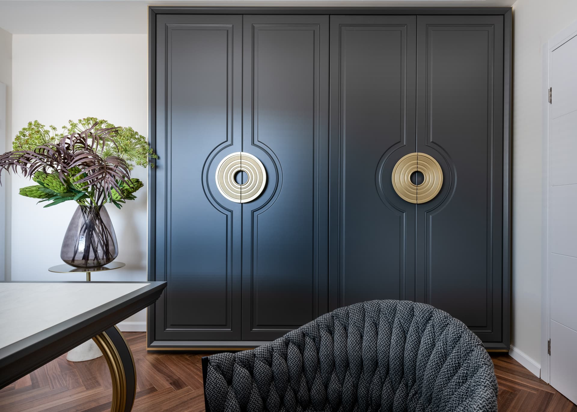 Dark panelled wardrobe with circular brass disc handles and a woven armchair in the consultation office of Moeen Clinic