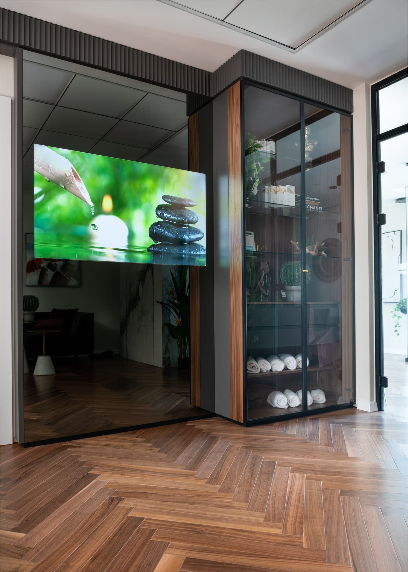 Walnut-framed glass cabinet with rolled towels beside a dark glass wall and mounted screen at the spa entrance of Moeen Clinic