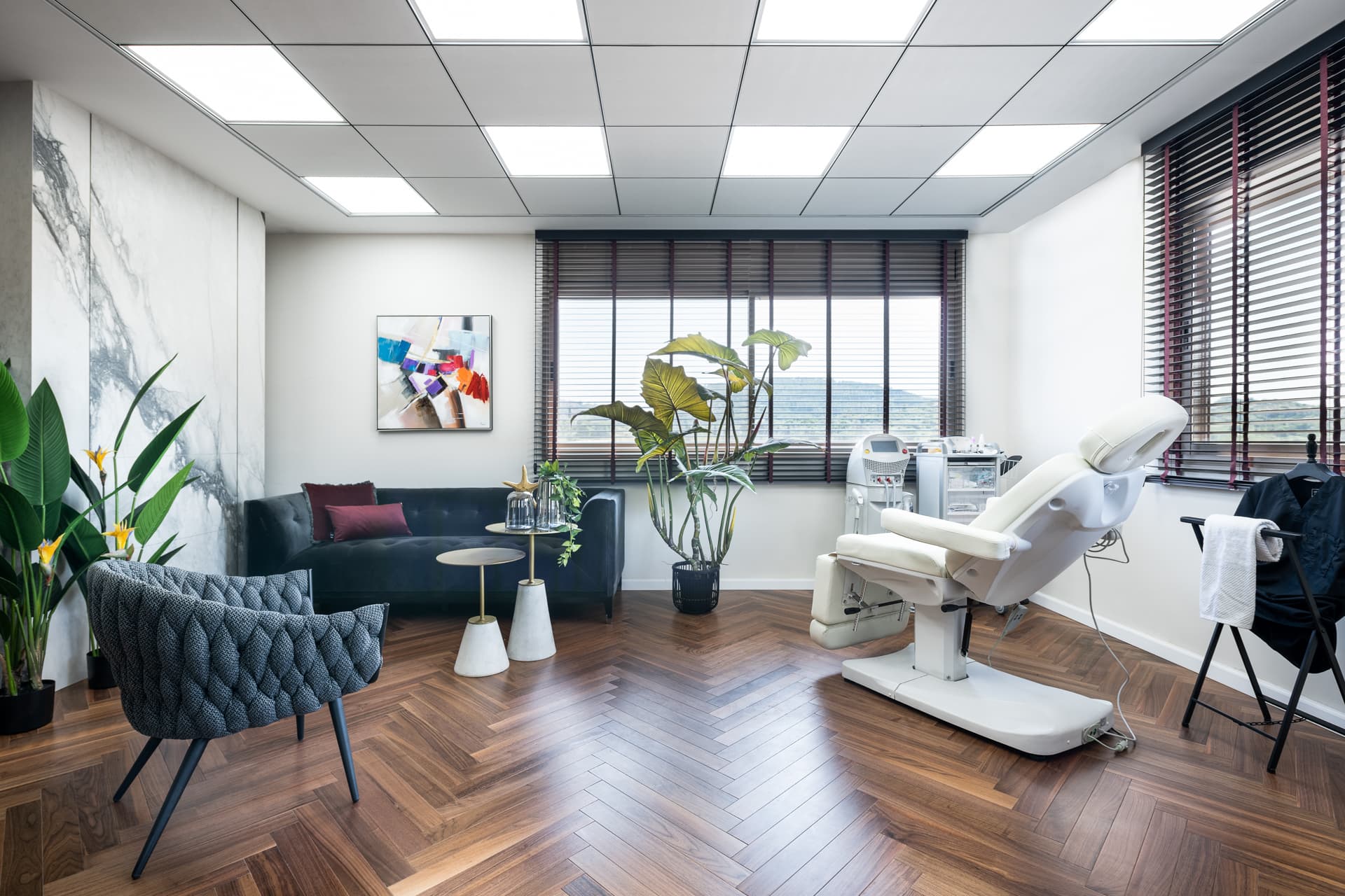Treatment chair beside a tufted velvet sofa, woven armchair and wooden blinds in the treatment room of Moeen Clinic