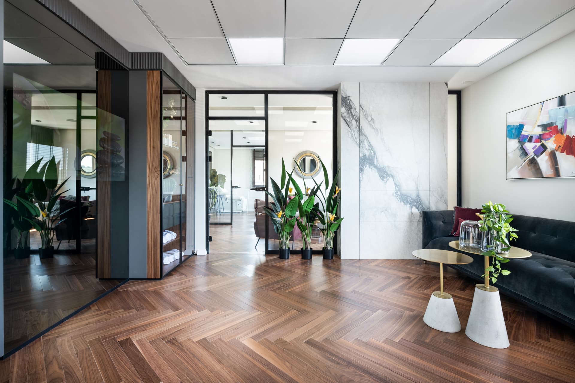 Seating area with glass partitions, a veined stone wall panel and brass-topped side tables in the treatment room of Moeen Clinic