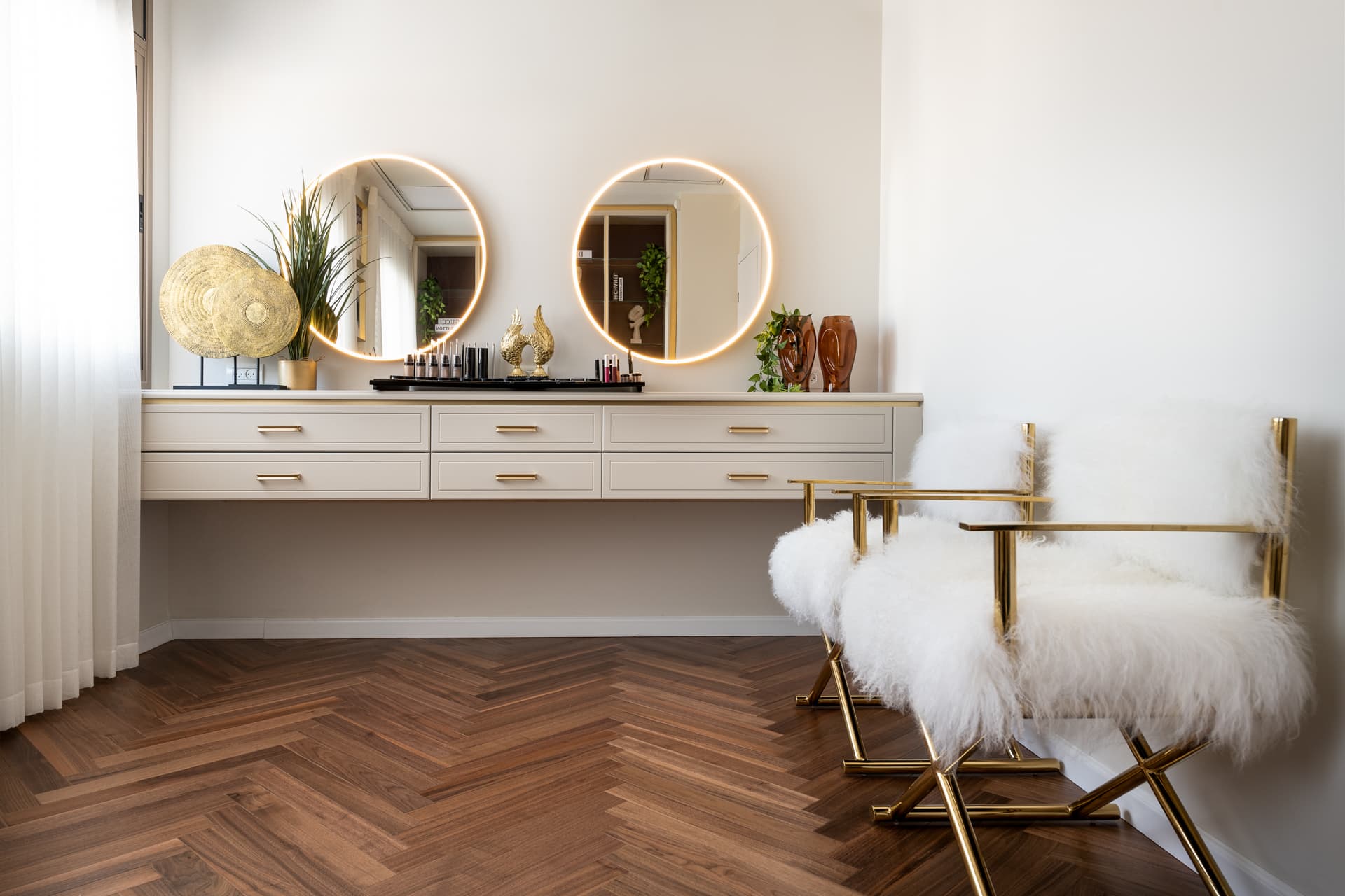 Floating cream vanity with brass pulls, two backlit round mirrors and gold-framed sheepskin chairs at the vanity station of Moeen Clinic