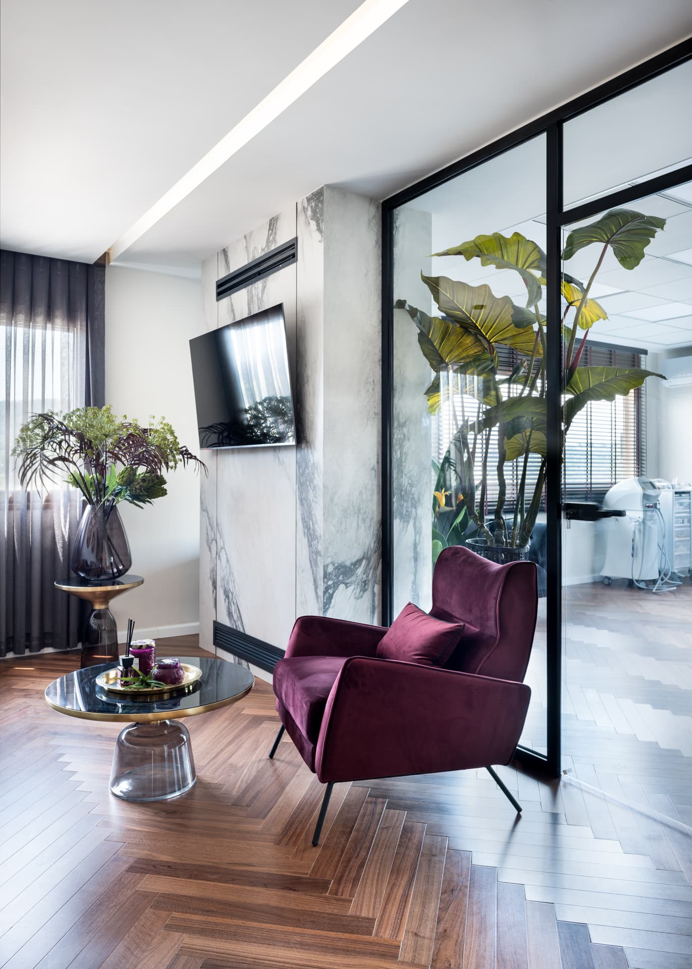 Plum velvet armchair and glass-based coffee table beside a veined stone TV wall and glass partition in the waiting lounge of Moeen Clinic