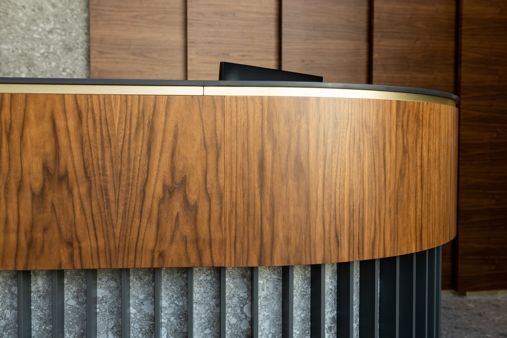 Close-up of the curved walnut reception desk front with a thin brass trim over dark fins and speckled stone