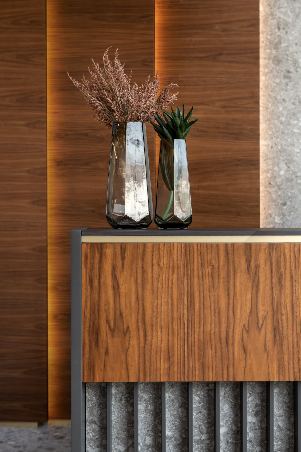 Faceted smoked glass vases with dried grasses on a walnut and brass-trimmed console against backlit walnut panels in the office reception