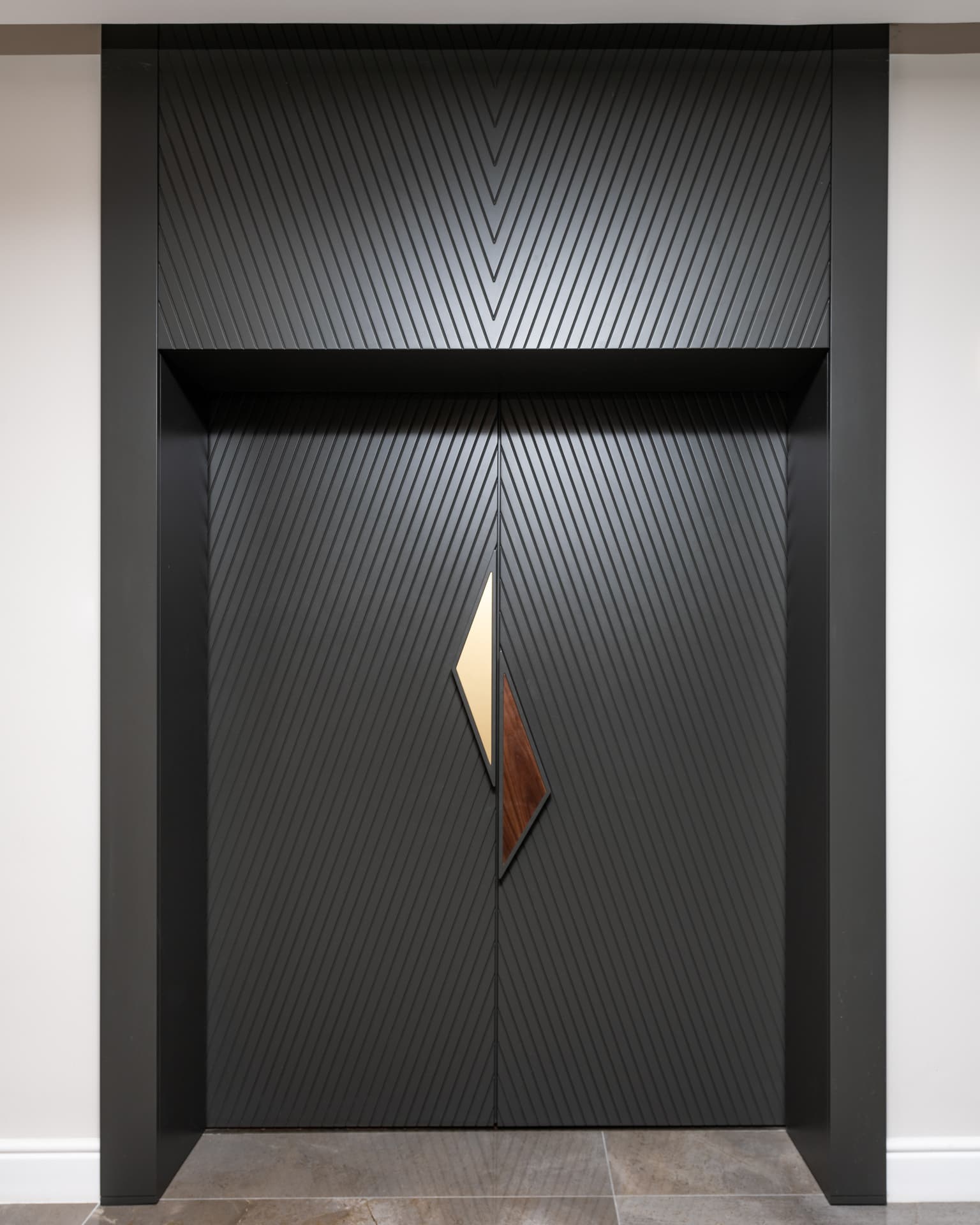 Frontal view of the chevron-grooved black entry doors and deep black portal frame with a brass and walnut pull at the office entrance
