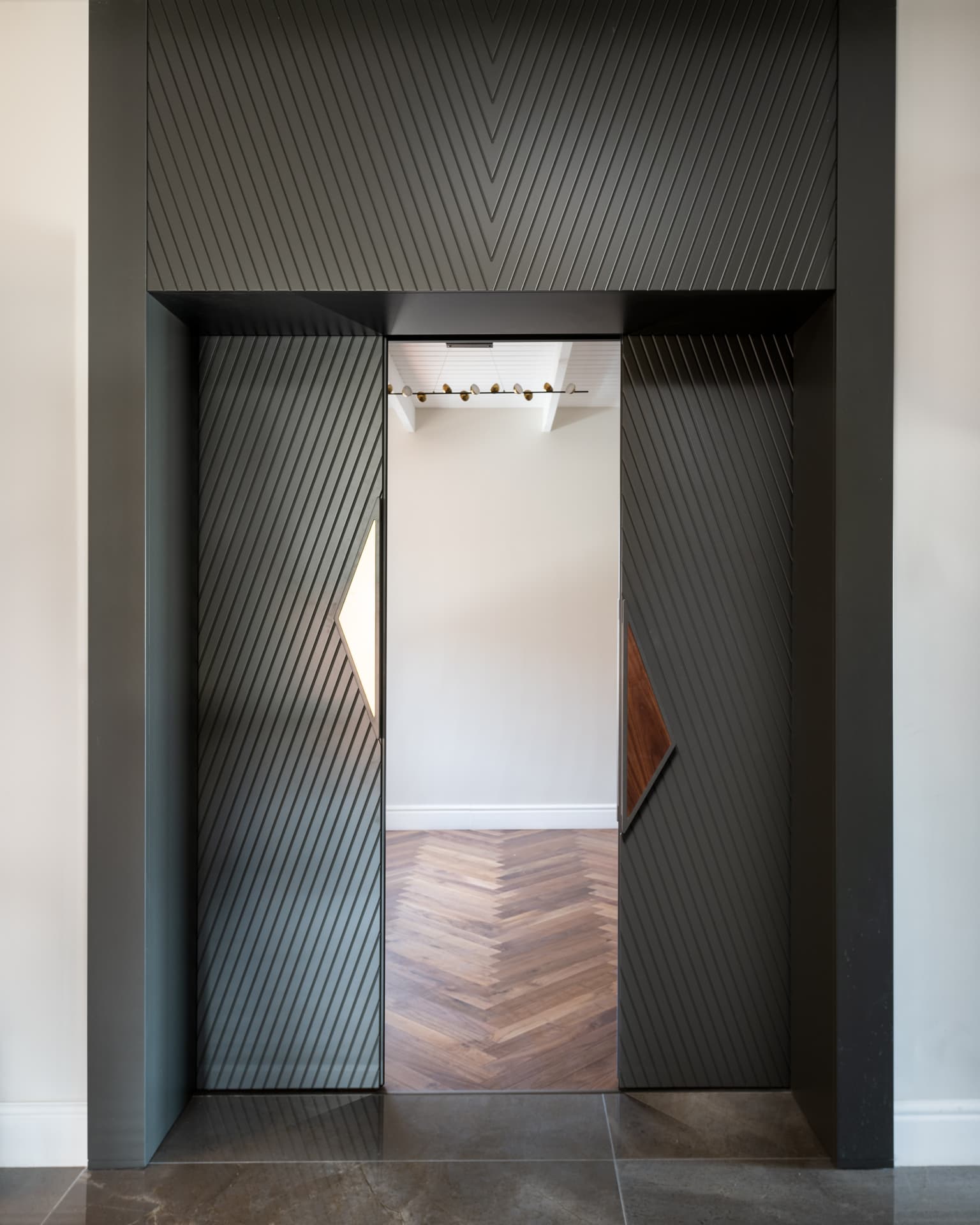 Grooved black entry doors opened onto a room with herringbone wood flooring and a brass rail light in the office entrance