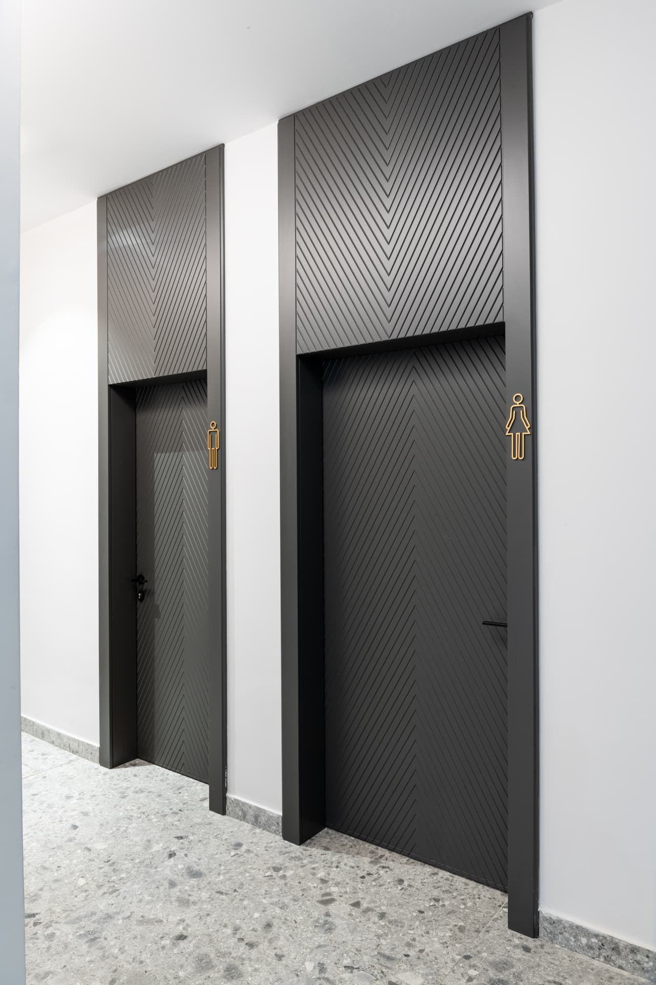 Two tall chevron-grooved charcoal doors with brass restroom pictograms along a terrazzo corridor near the office entrance