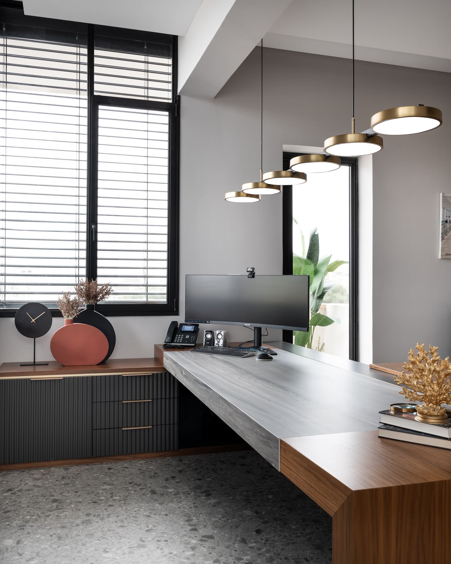 Cantilevered veined stone desk meeting a walnut block under a stepped row of brass disc pendants in the executive office