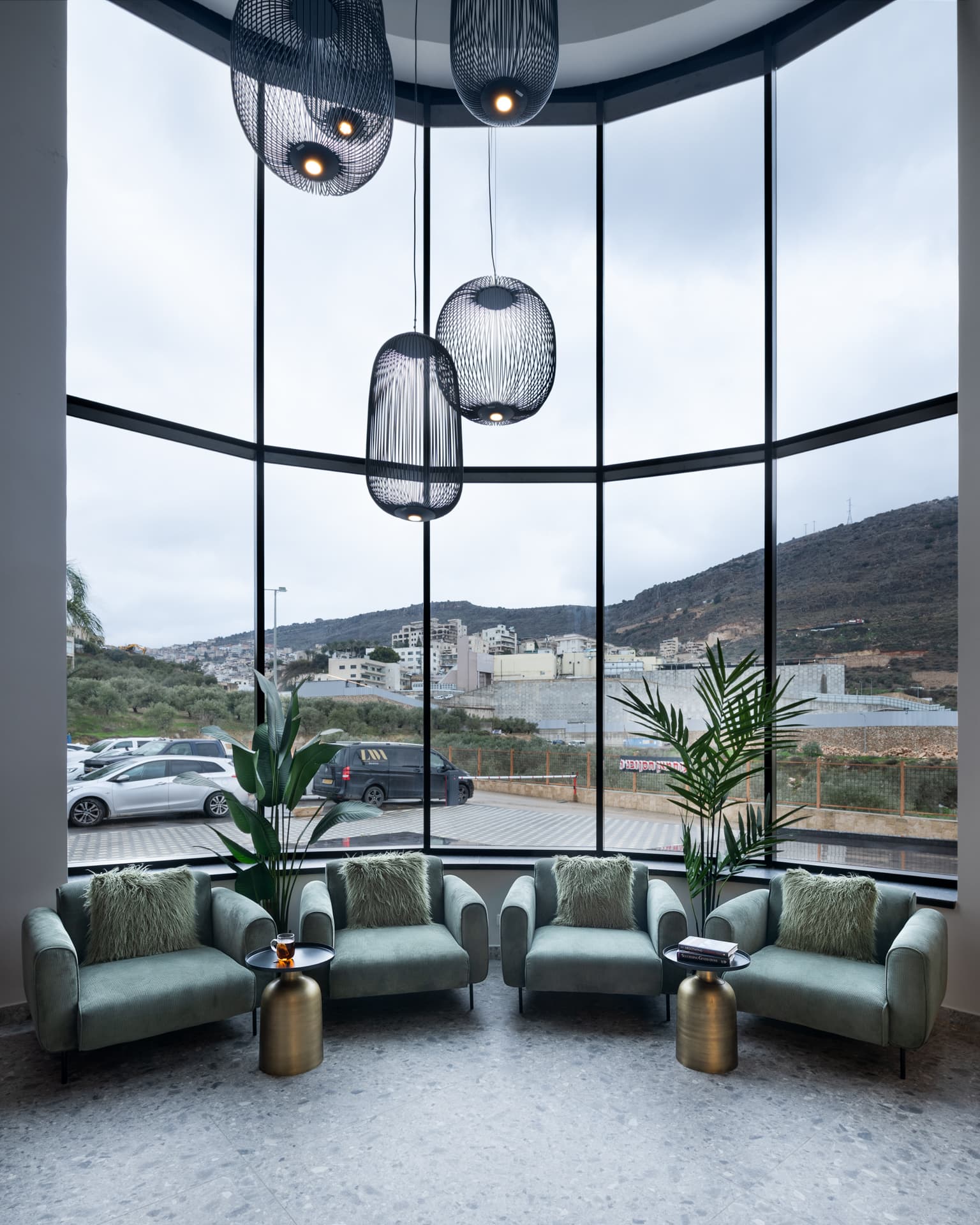 Four sage velvet armchairs with shaggy cushions before a curved double-height window under black wire pendants in the office lounge
