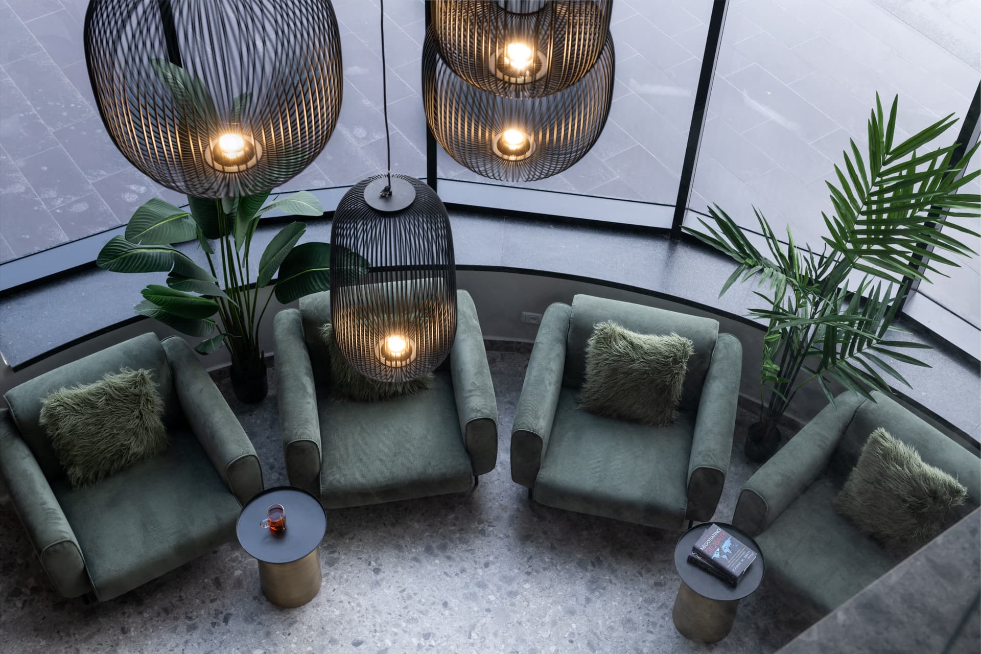 Overhead view of sage velvet armchairs and brass drum side tables beneath glowing black wire cage pendants in the office lounge