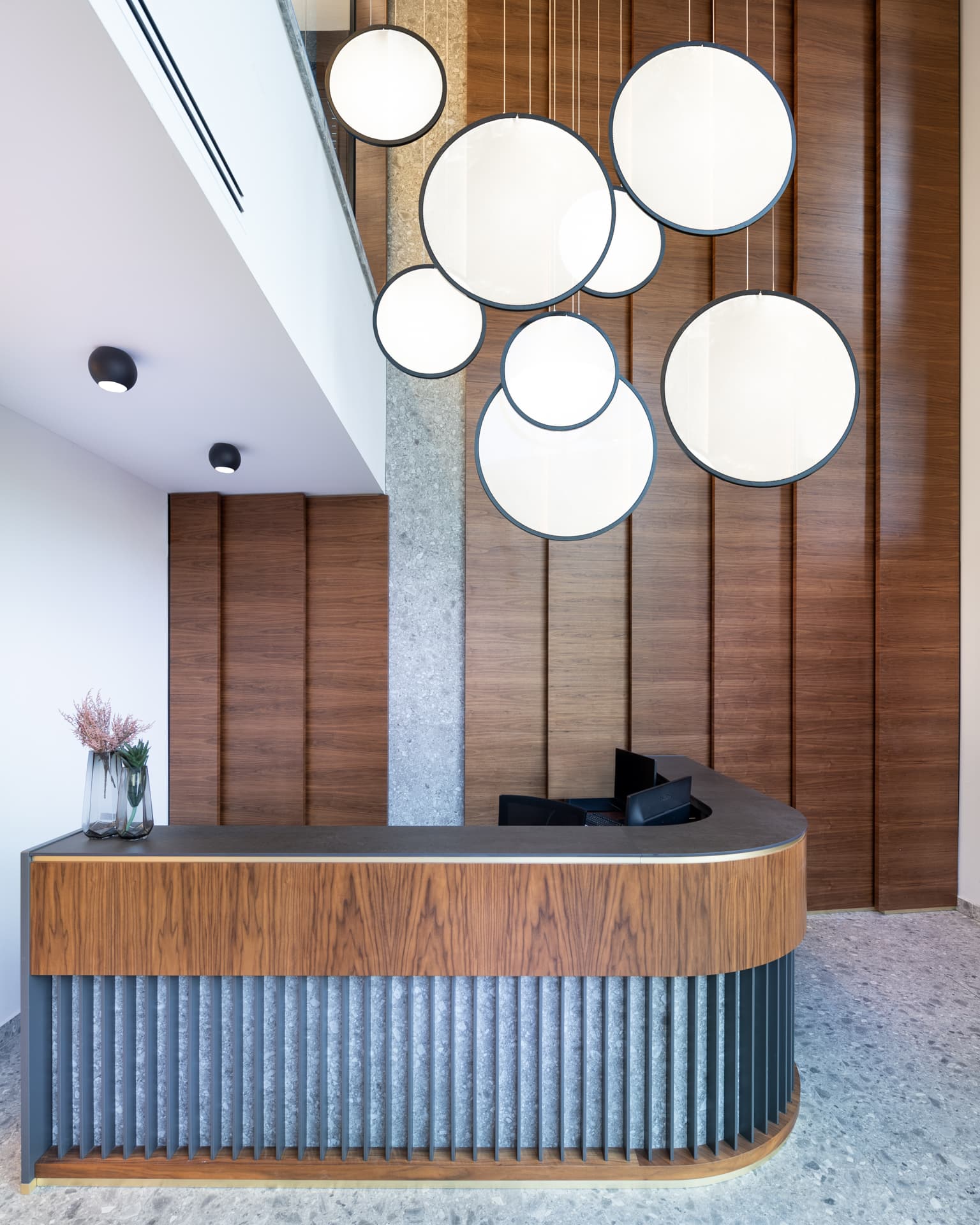 Curved walnut reception desk with dark vertical fins beneath a cluster of glowing disc pendants in the office reception