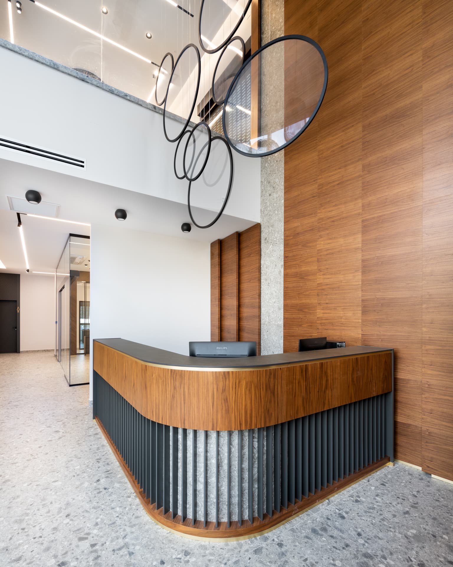 Angled view of the curved walnut reception desk and full-height walnut wall under black ring pendants in the office reception