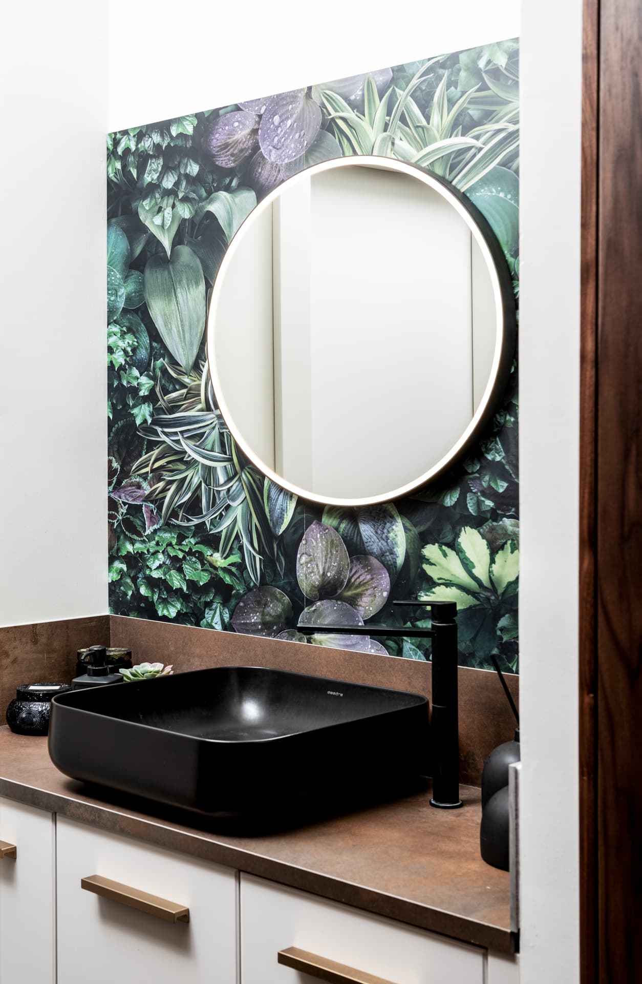 Black vessel basin and matte black faucet below a round backlit mirror on a leafy botanical wall in the bathroom of Othman Office 2