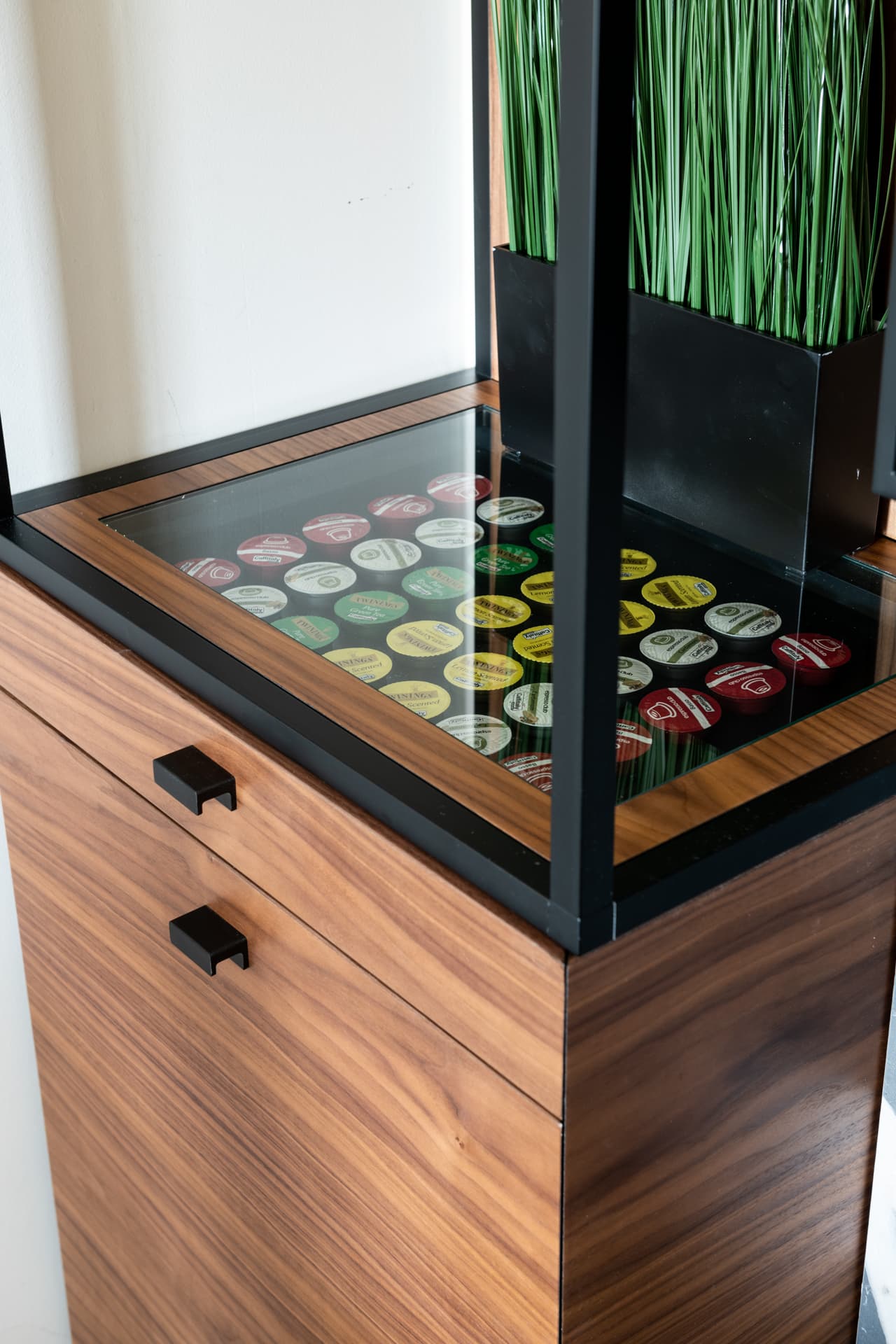 Glass-topped walnut cabinet showing rows of coffee capsules below a grass planter in a steel frame in the conference room of Othman Office 2