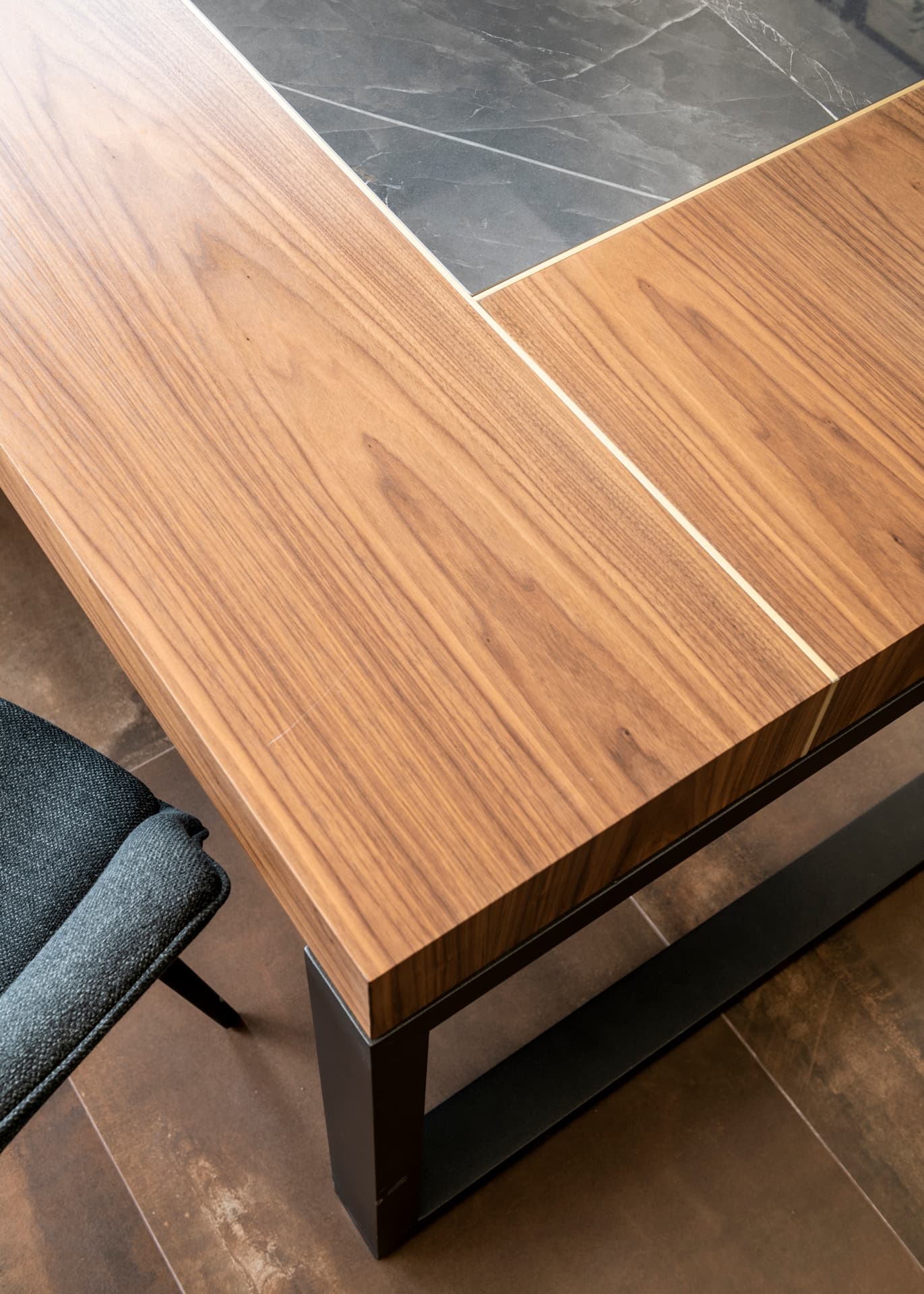 Corner of a walnut desktop with an inset dark stone panel and a thin brass seam over a steel leg in the private office of Othman Office 2
