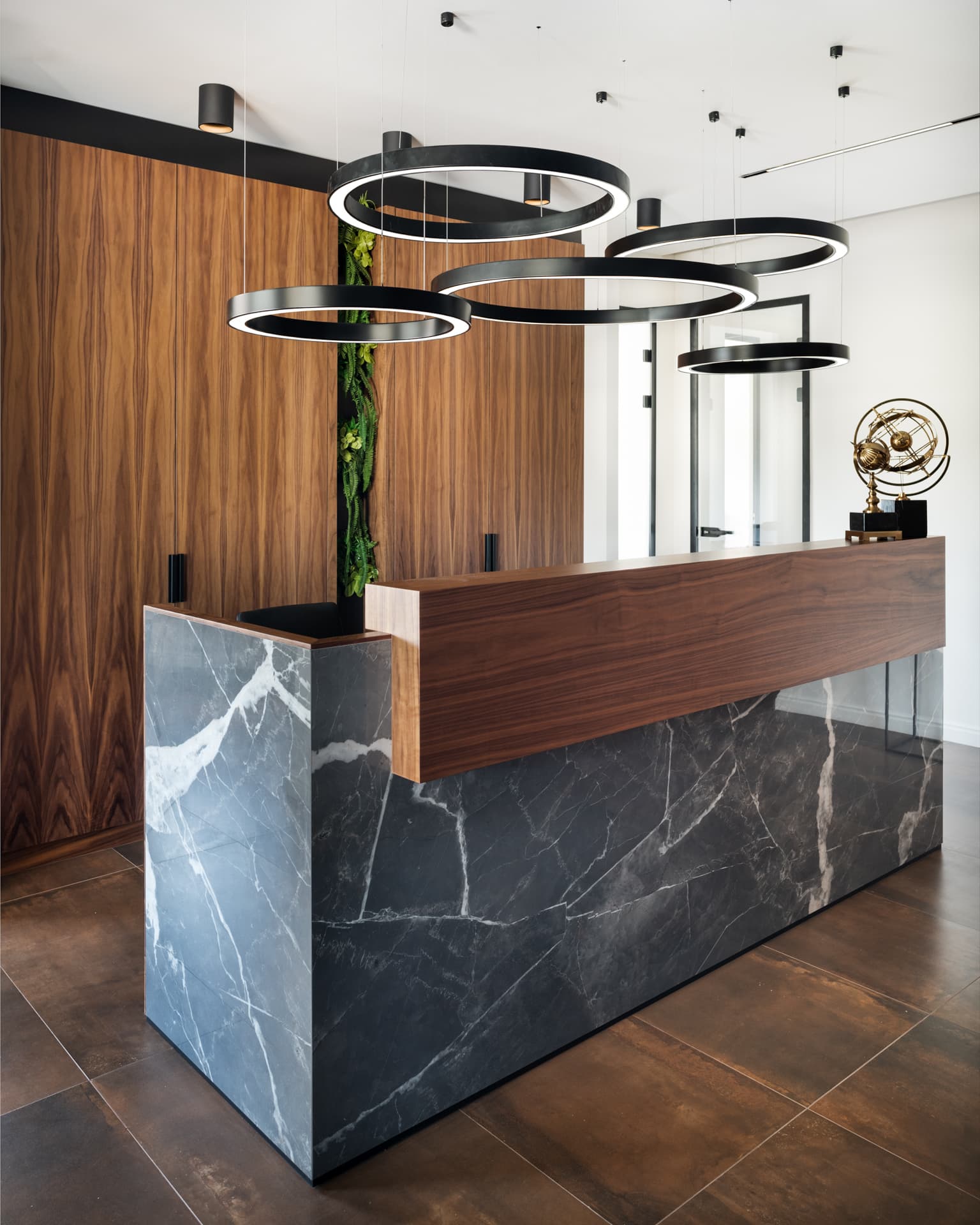 Corner view of the dark veined stone reception desk with a floating walnut top below black ring pendants in the reception of Othman Office 2