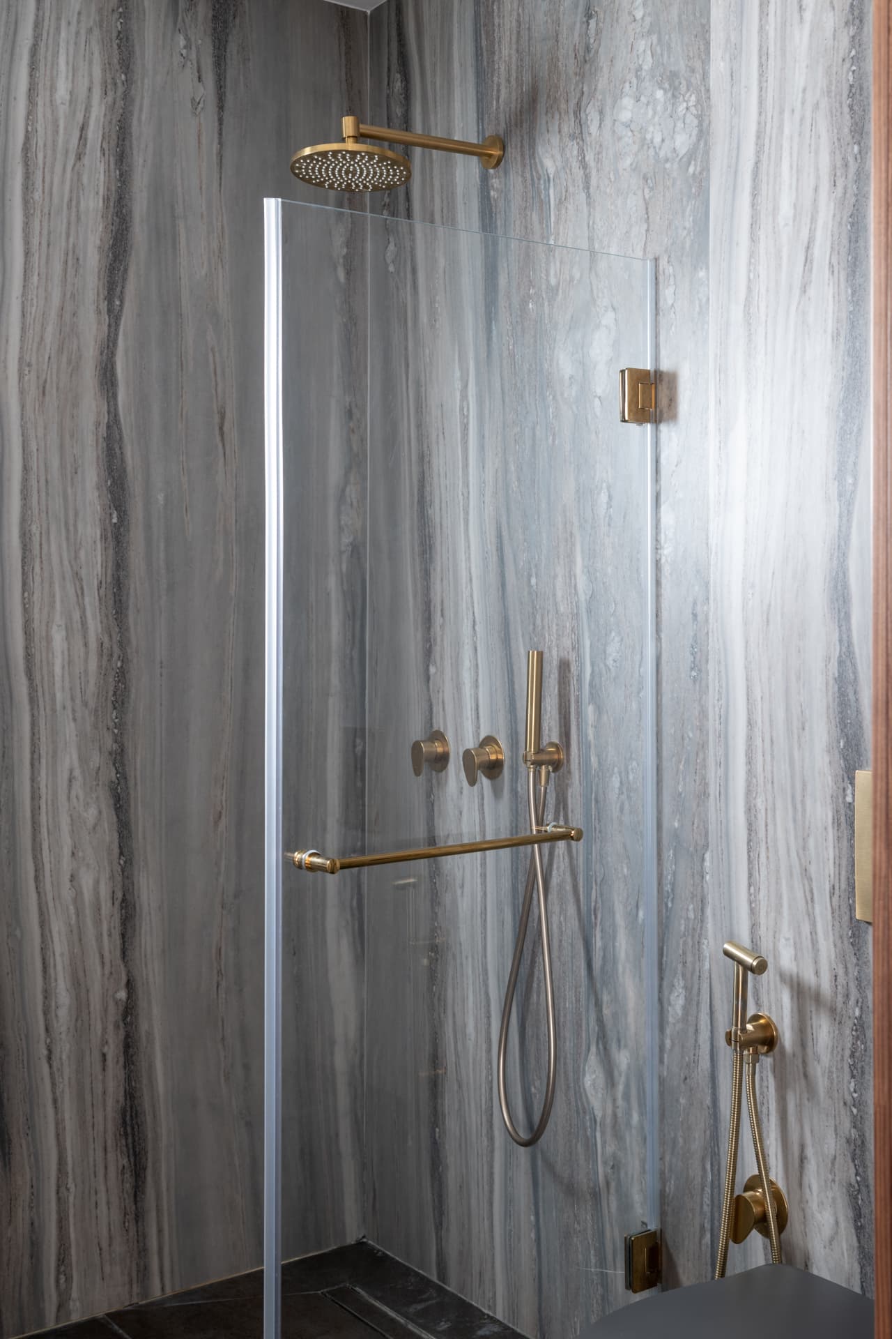 Walk-in shower with brushed brass rain head and fittings behind a glass panel on grey veined stone in the master bathroom of Pistachio