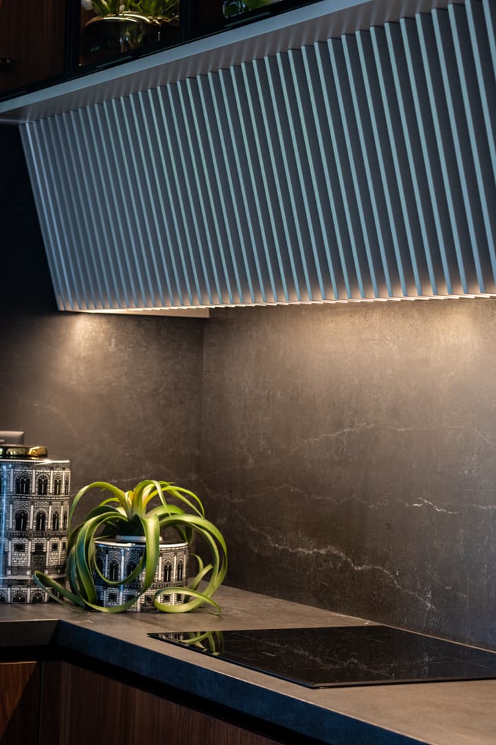 Fluted angled hood panel lit from below over a dark veined stone backsplash and black induction hob in the kitchen of Pistachio