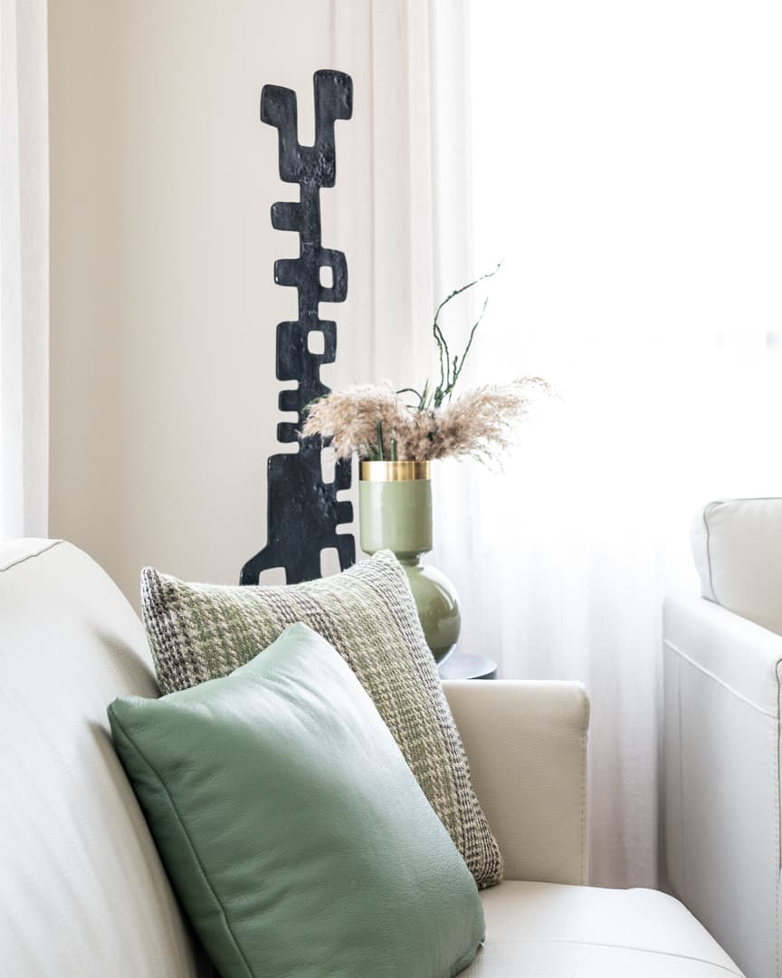 Sage leather and woven cushions on a cream sofa before a black totem sculpture and brass-rimmed green vase in the living room of Pistachio