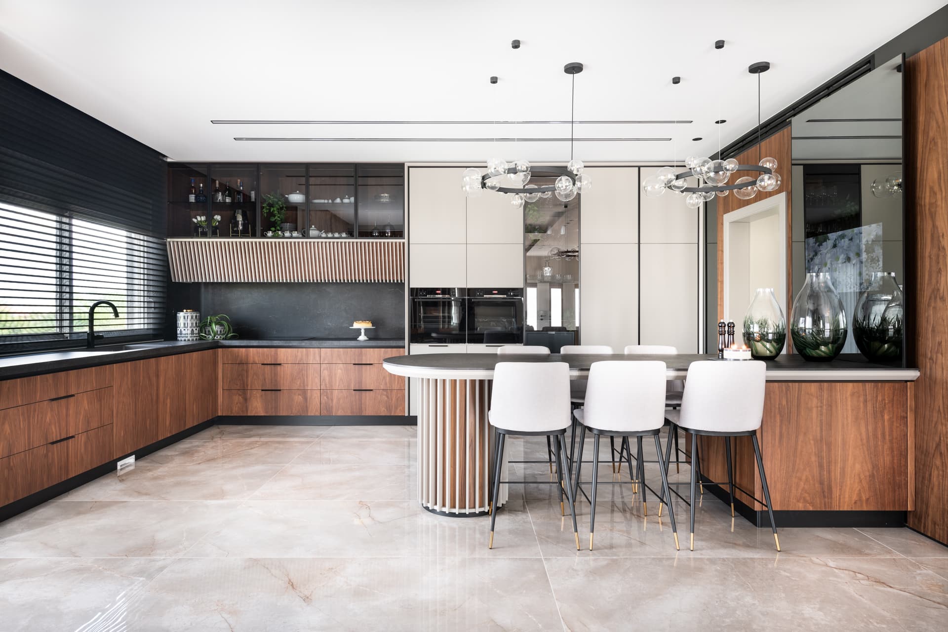 Walnut kitchen with dark stone counters, lit glass-front cabinets and a curved island with pale stools in the kitchen of Pistachio