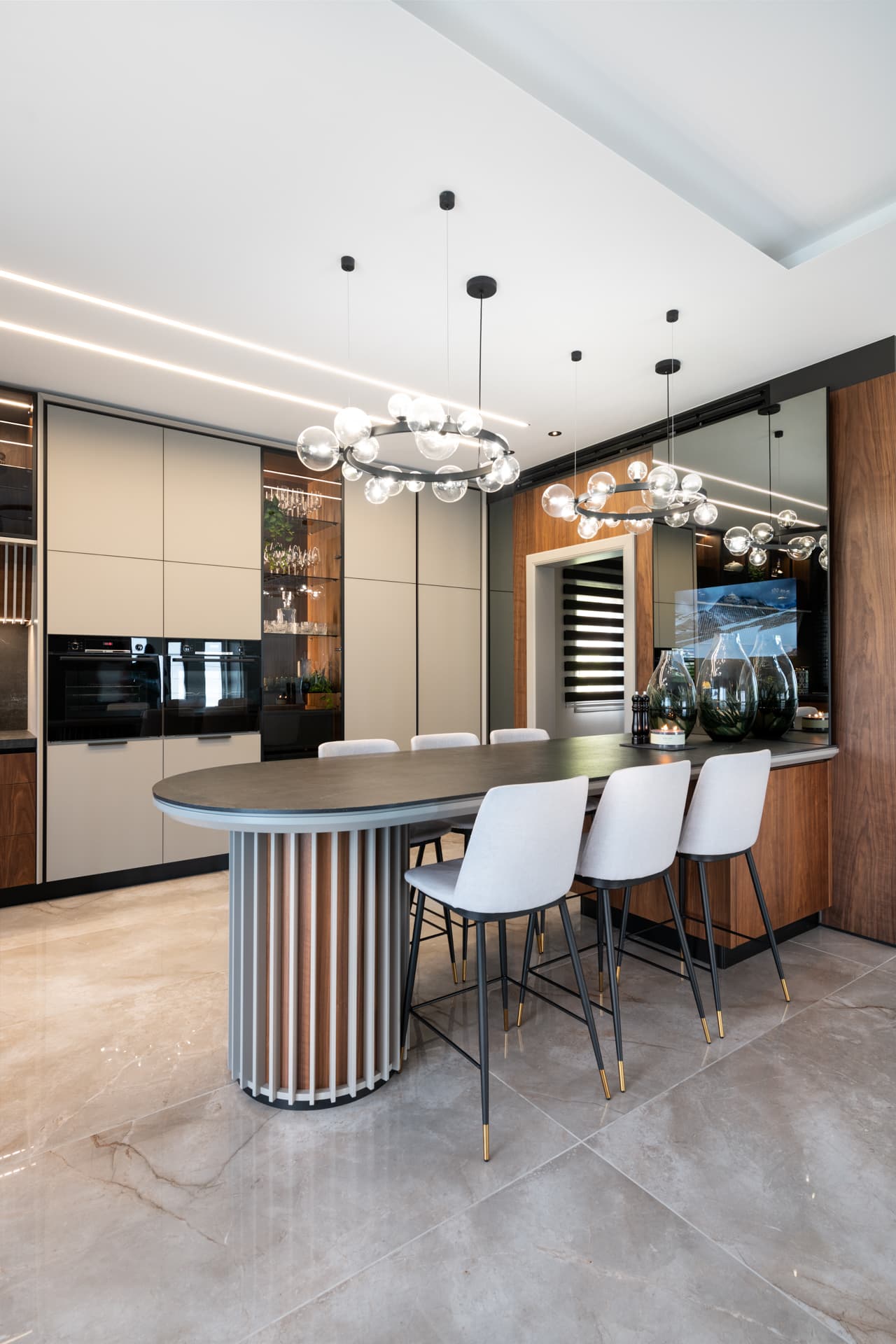 Oval island on a slatted round base under two glass-globe ring chandeliers, with tall built-in ovens in the kitchen of Pistachio