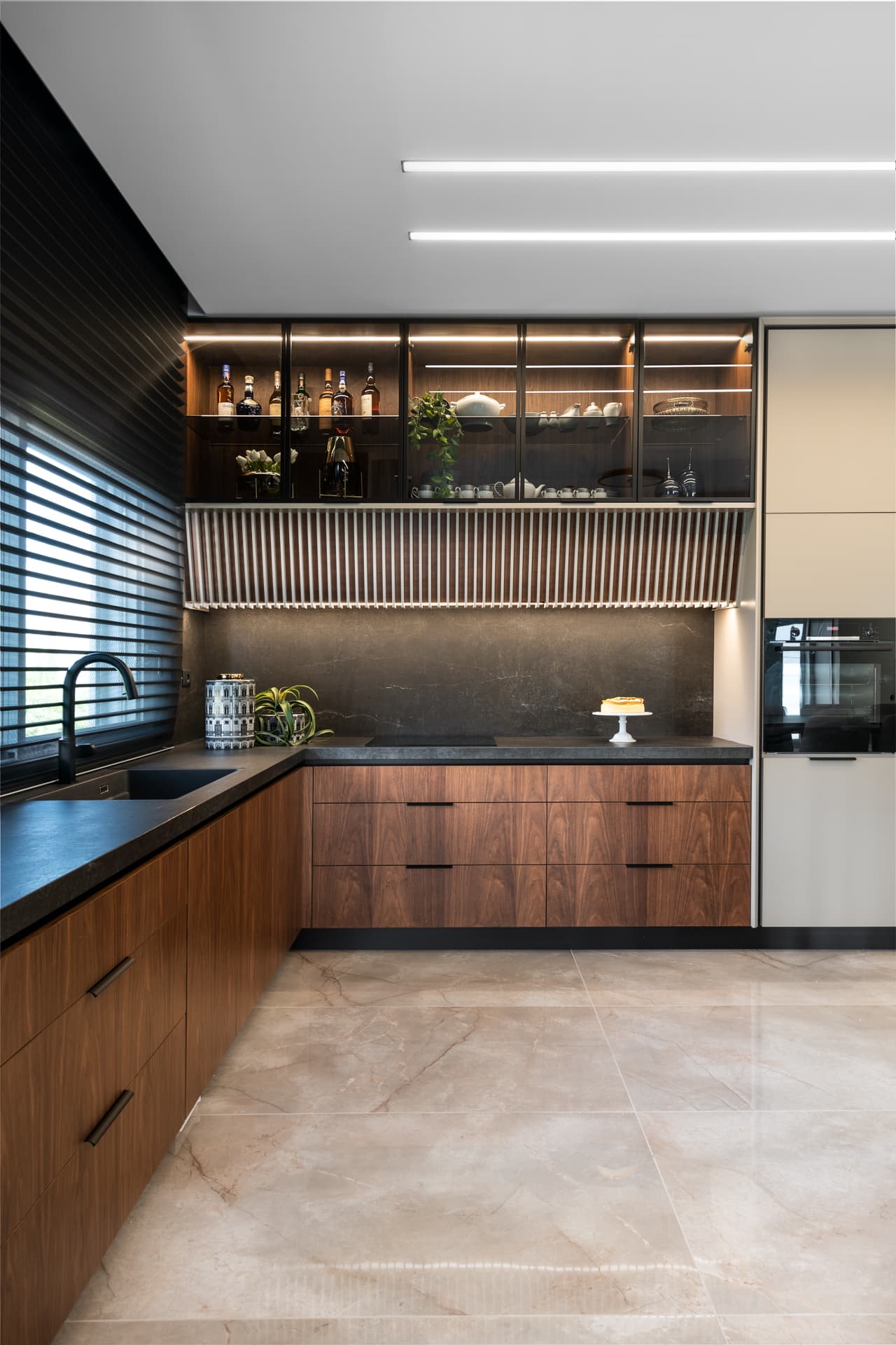 Corner of walnut drawers, dark veined stone backsplash and lit glass-front upper shelving over a fluted panel in the kitchen of Pistachio