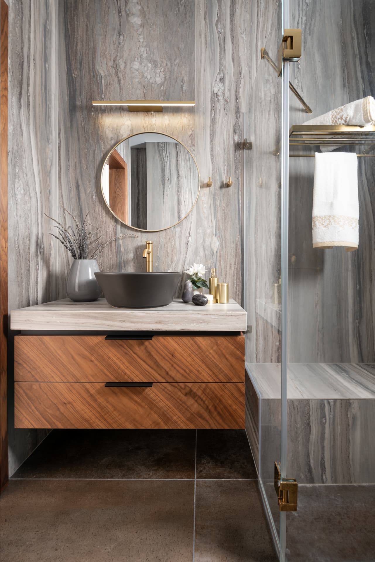 Dark vessel basin on a walnut vanity against grey veined stone walls, with a round brass mirror in the master bathroom of Pistachio