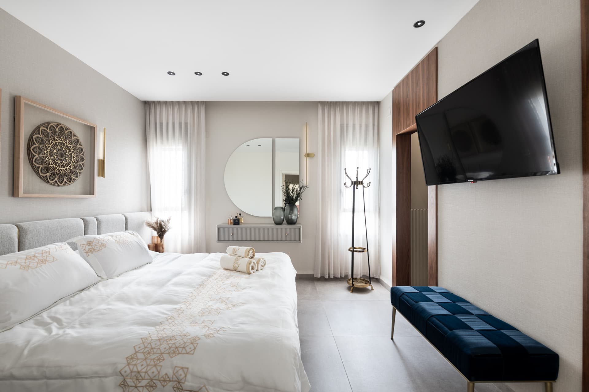 Upholstered grey panel headboard, framed mandala art and a navy velvet bench under a wall-mounted screen in the master bedroom of Pistachio