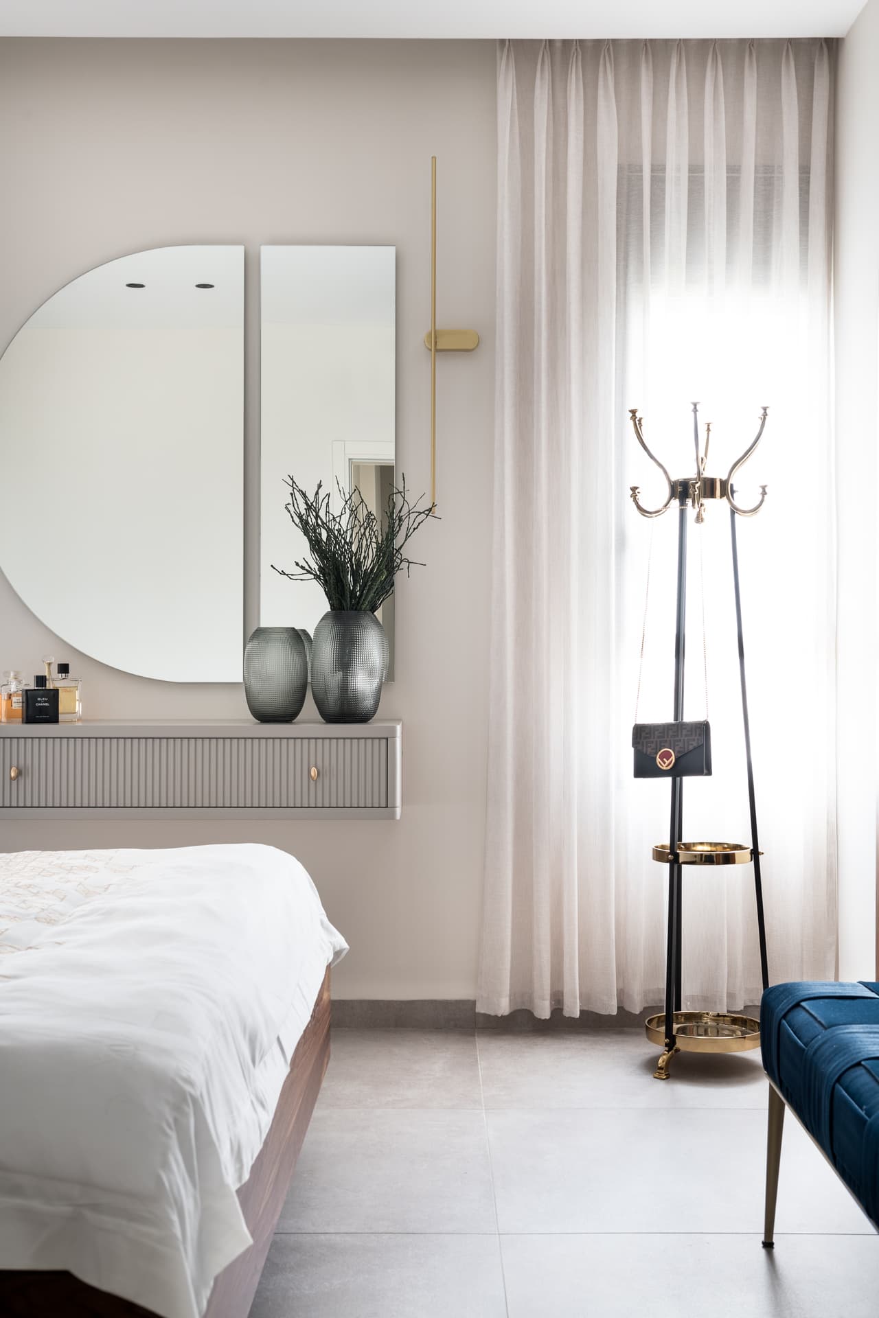 Split round mirror over a fluted floating vanity, beside a brass-topped standing coat rack at the window in the master bedroom of Pistachio