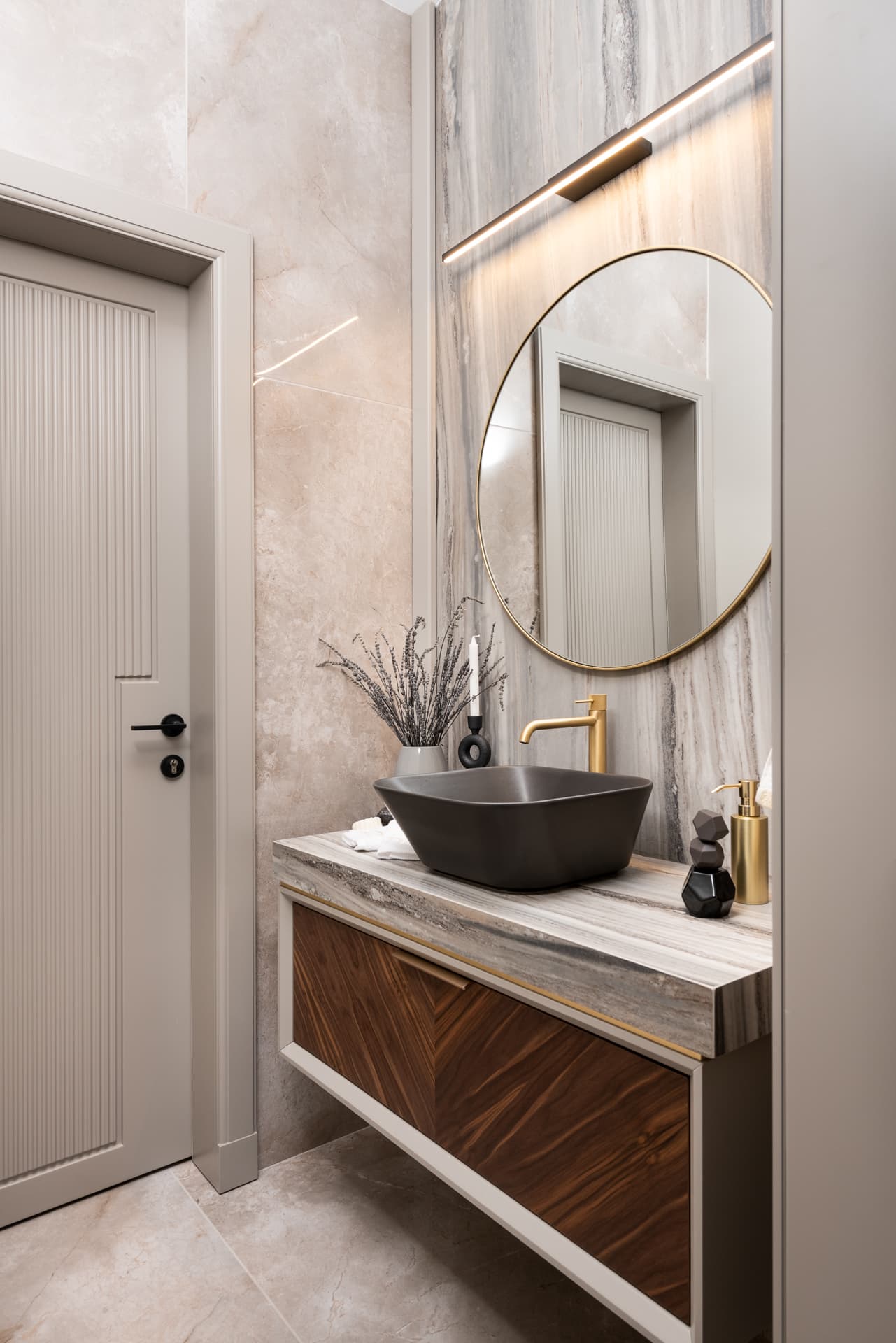 Dark square vessel basin on a walnut vanity with a stone top, under a round brass mirror and linear light in the powder room of Pistachio