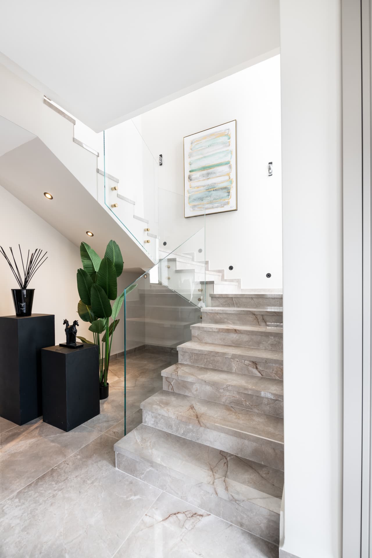 Beige veined stone staircase with a frameless glass balustrade, black plinths and a tall potted plant at its base in Pistachio