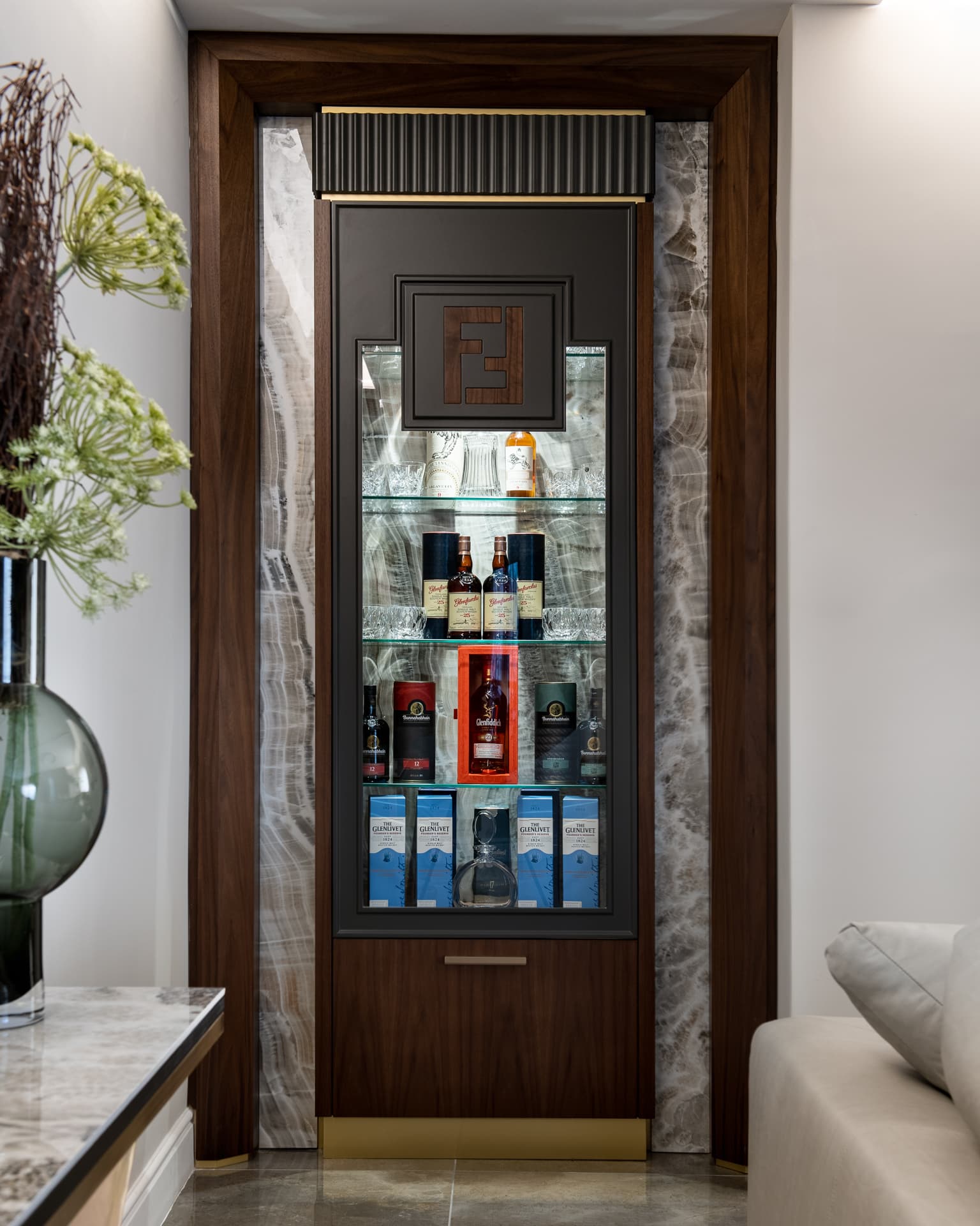 Walnut-framed glass display cabinet flanked by backlit onyx panels and topped with fluted black trim in the bar of Saffron