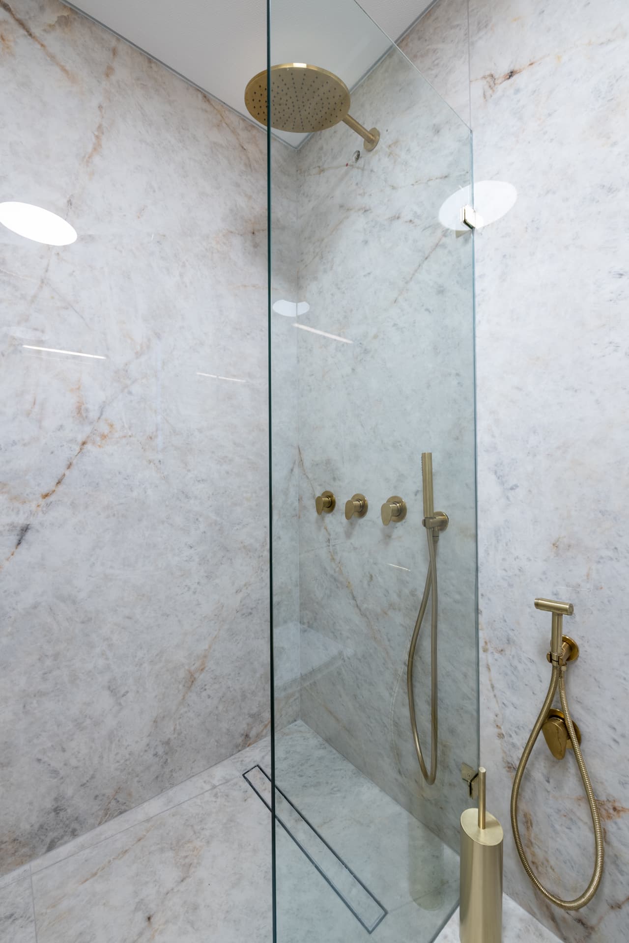 Frameless glass shower lined in veined stone with a brushed brass rain head, hand shower and linear floor drain in a bathroom of Saffron