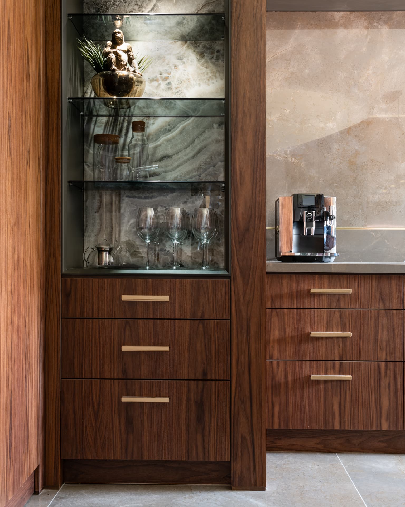 Walnut drawers with brass pulls below a lit glass-shelf niche backed with veined stone in the kitchen of Saffron