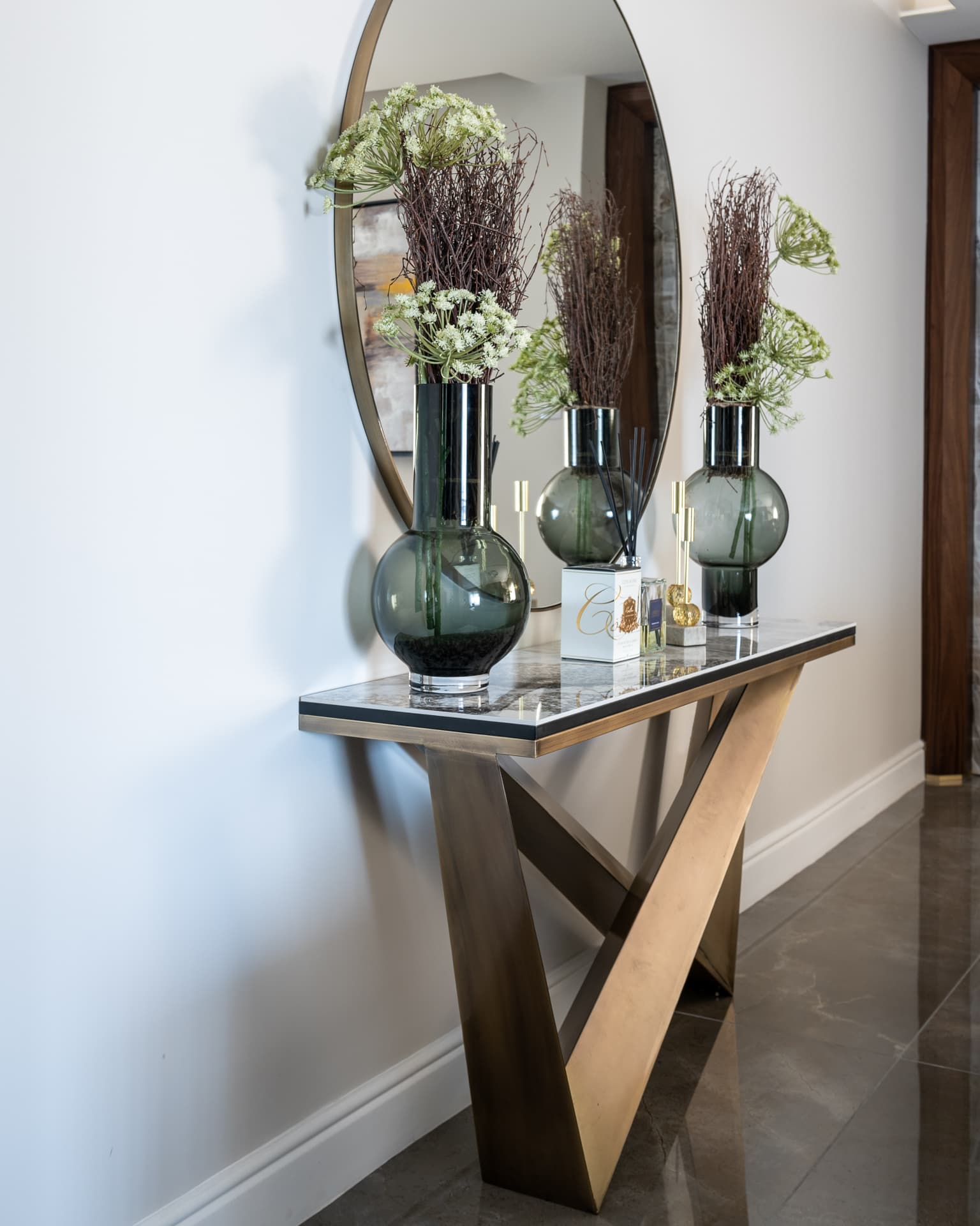 Angular bronze console with a stone top, smoked-glass vases and a round brass-framed mirror in the entryway of Saffron