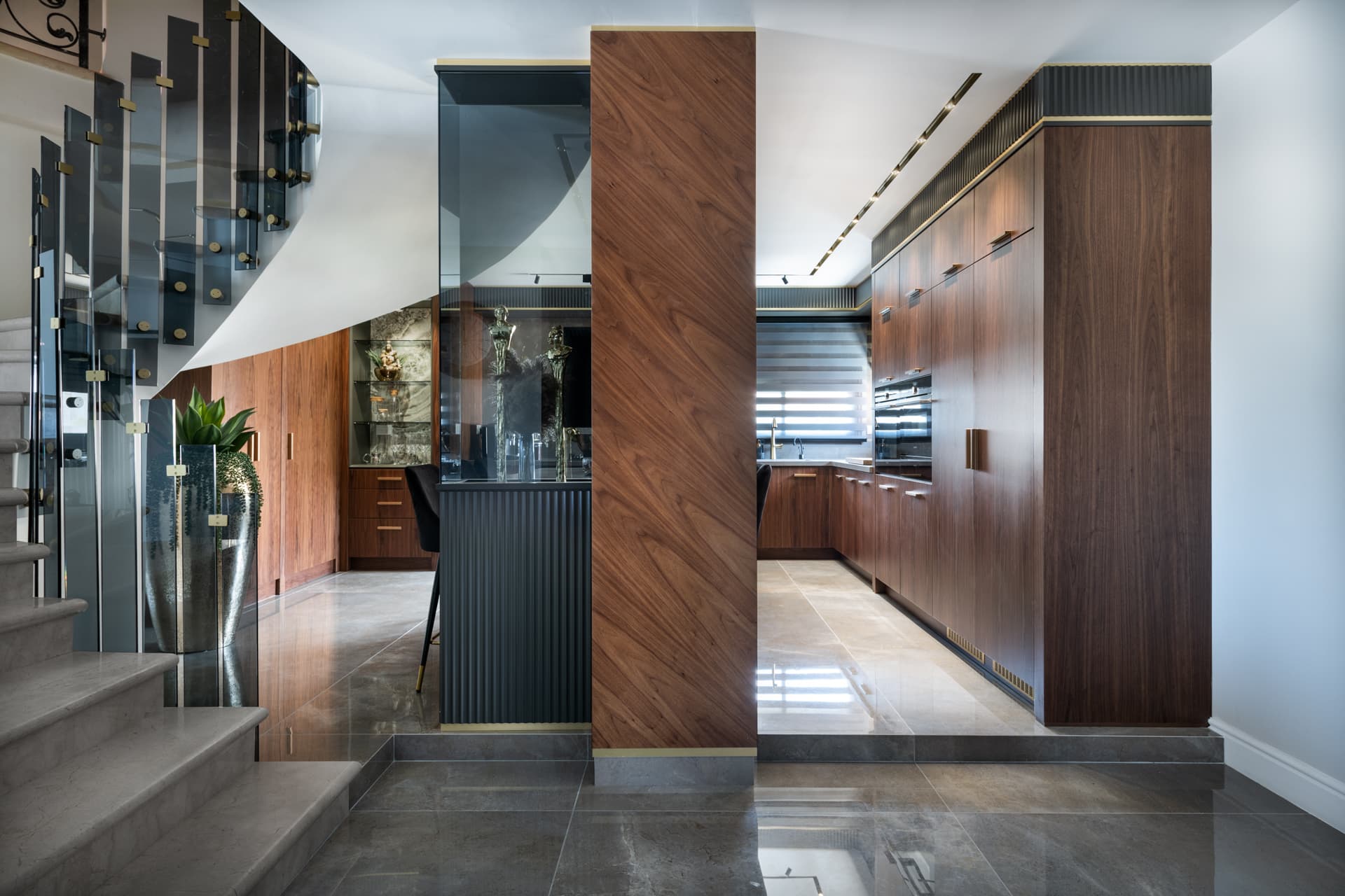Entry view past a smoked-glass staircase balustrade to a diagonal-grain walnut column and the walnut kitchen in Saffron