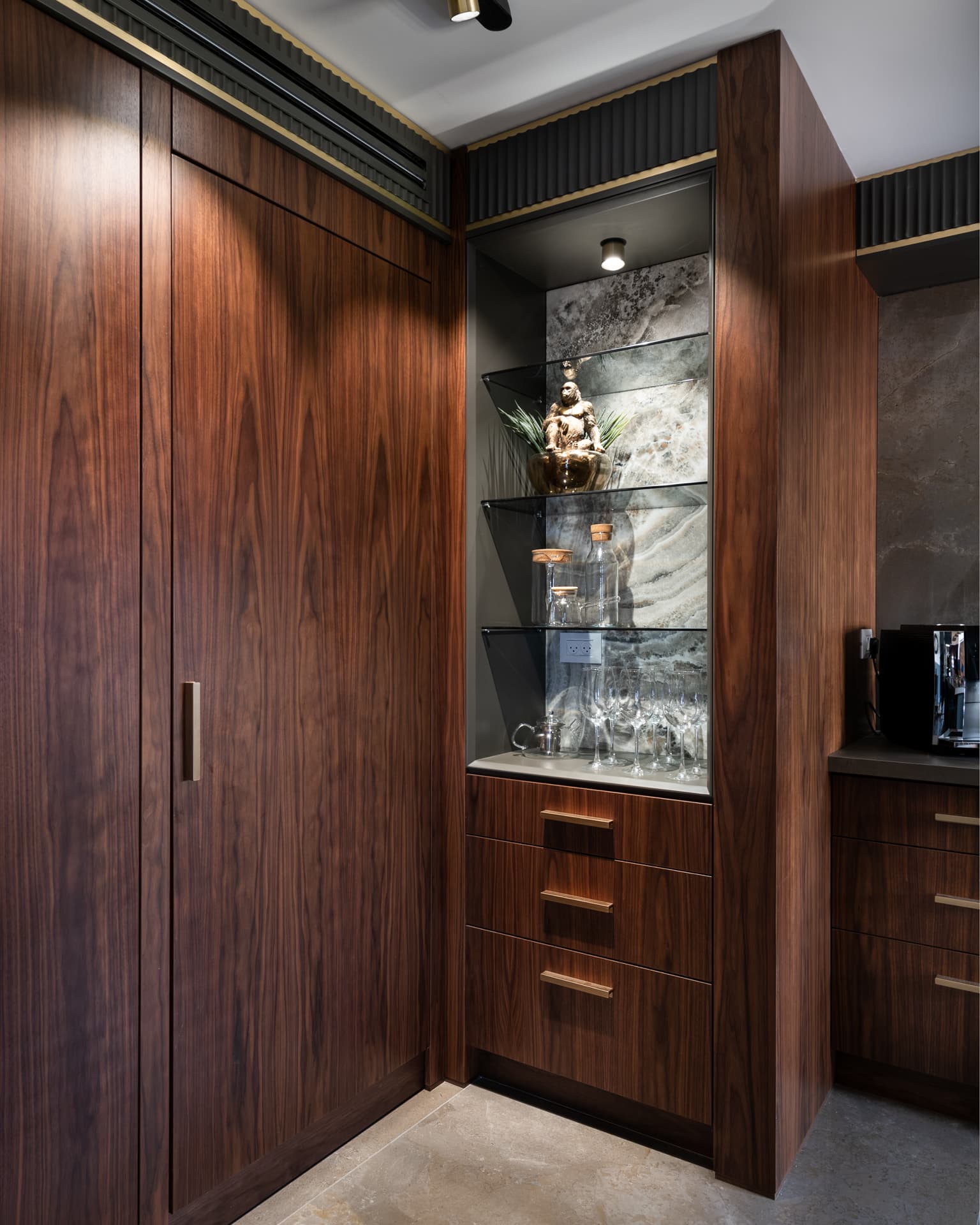 Tall walnut cabinetry beside a lit glass-shelf niche backed with veined stone and fluted black trim in the kitchen of Saffron