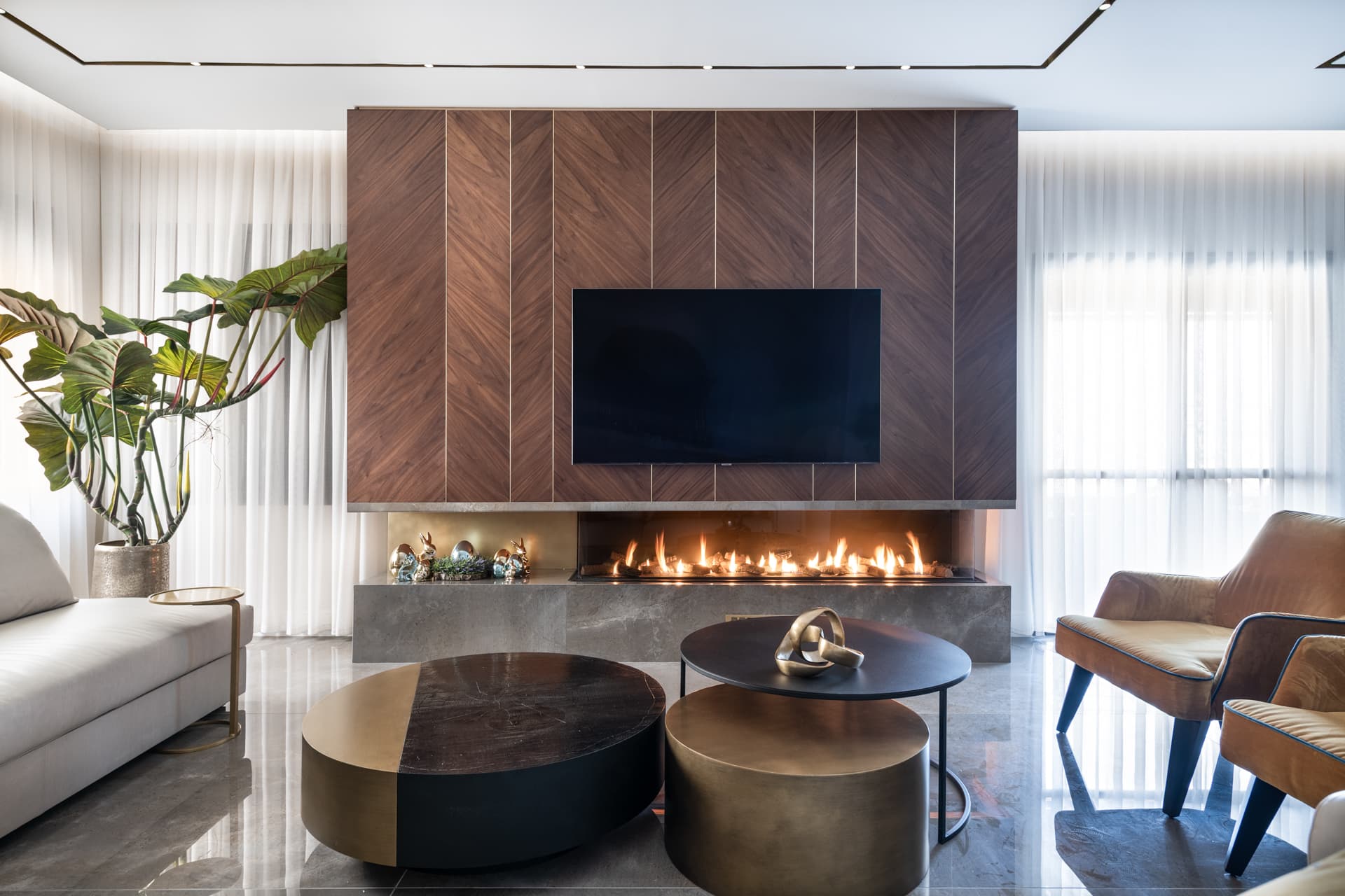 Chevron walnut media wall above a stone-clad linear fireplace, with round bronze and black coffee tables in the living room of Saffron