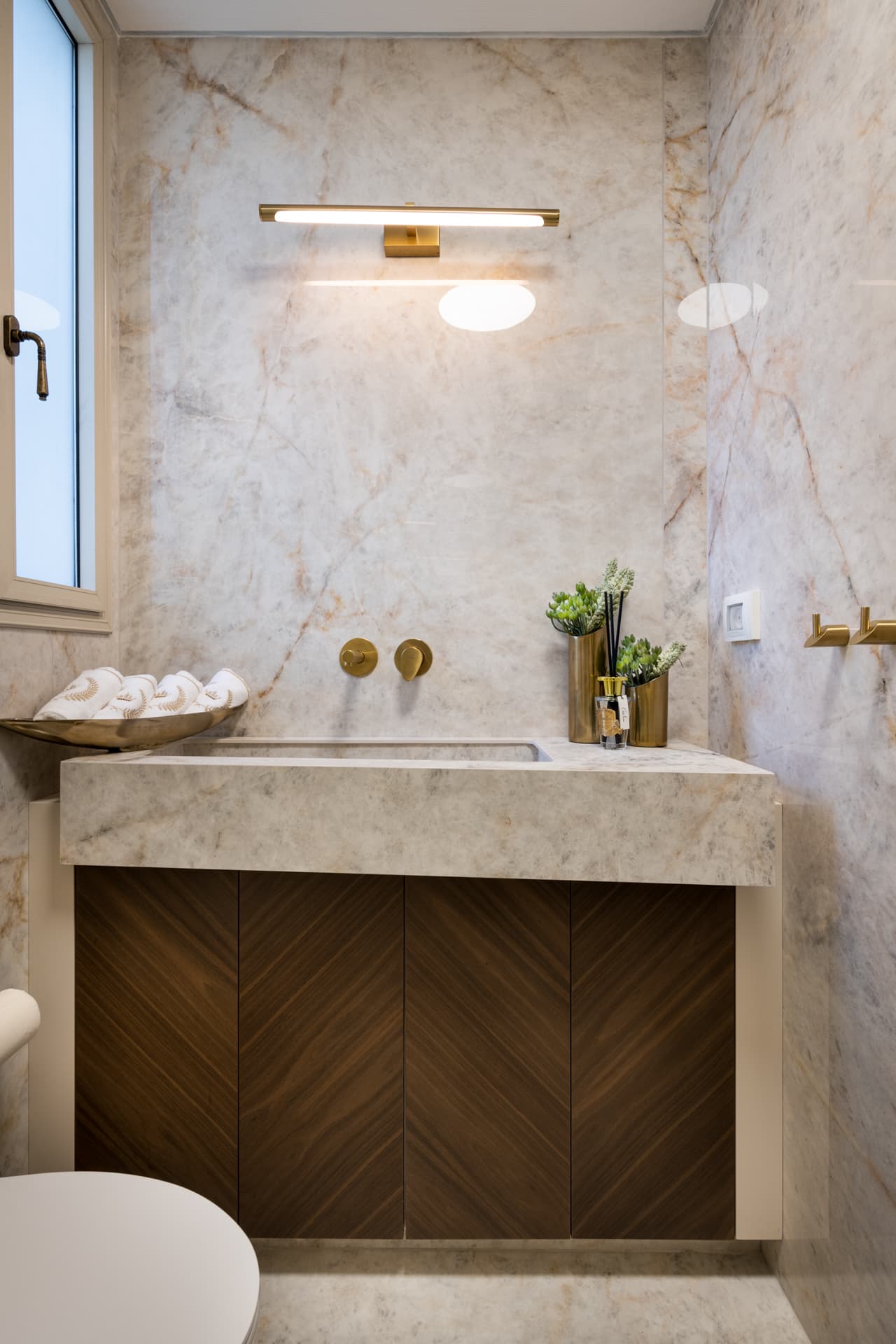 Veined stone sink and walls with brass wall-mounted taps and a chevron walnut vanity in the powder room of Saffron