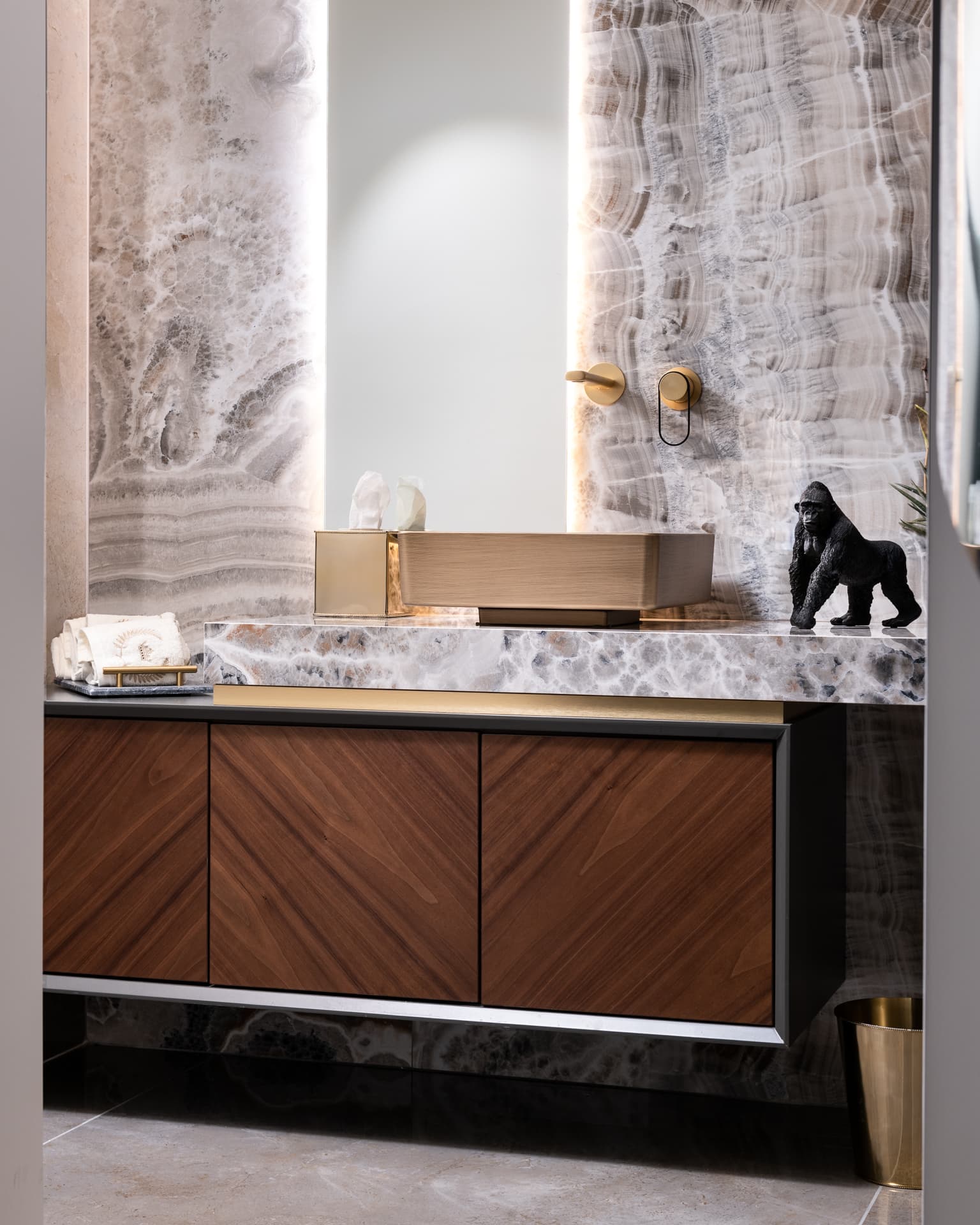Brass vessel basin on an onyx counter, backlit onyx walls and a chevron walnut vanity in the powder room of Saffron