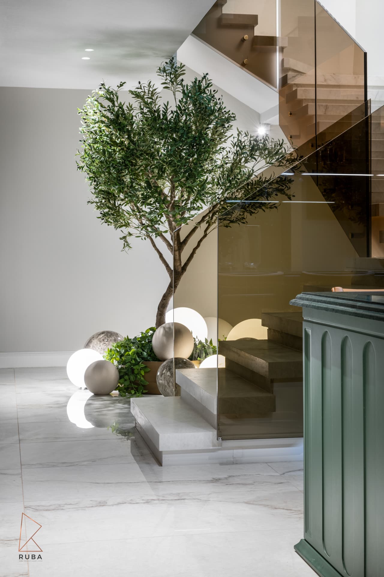 Stair void in the basement planted with an olive tree and glowing globe lights beside a stair with a bronze glass balustrade in The Last Supper