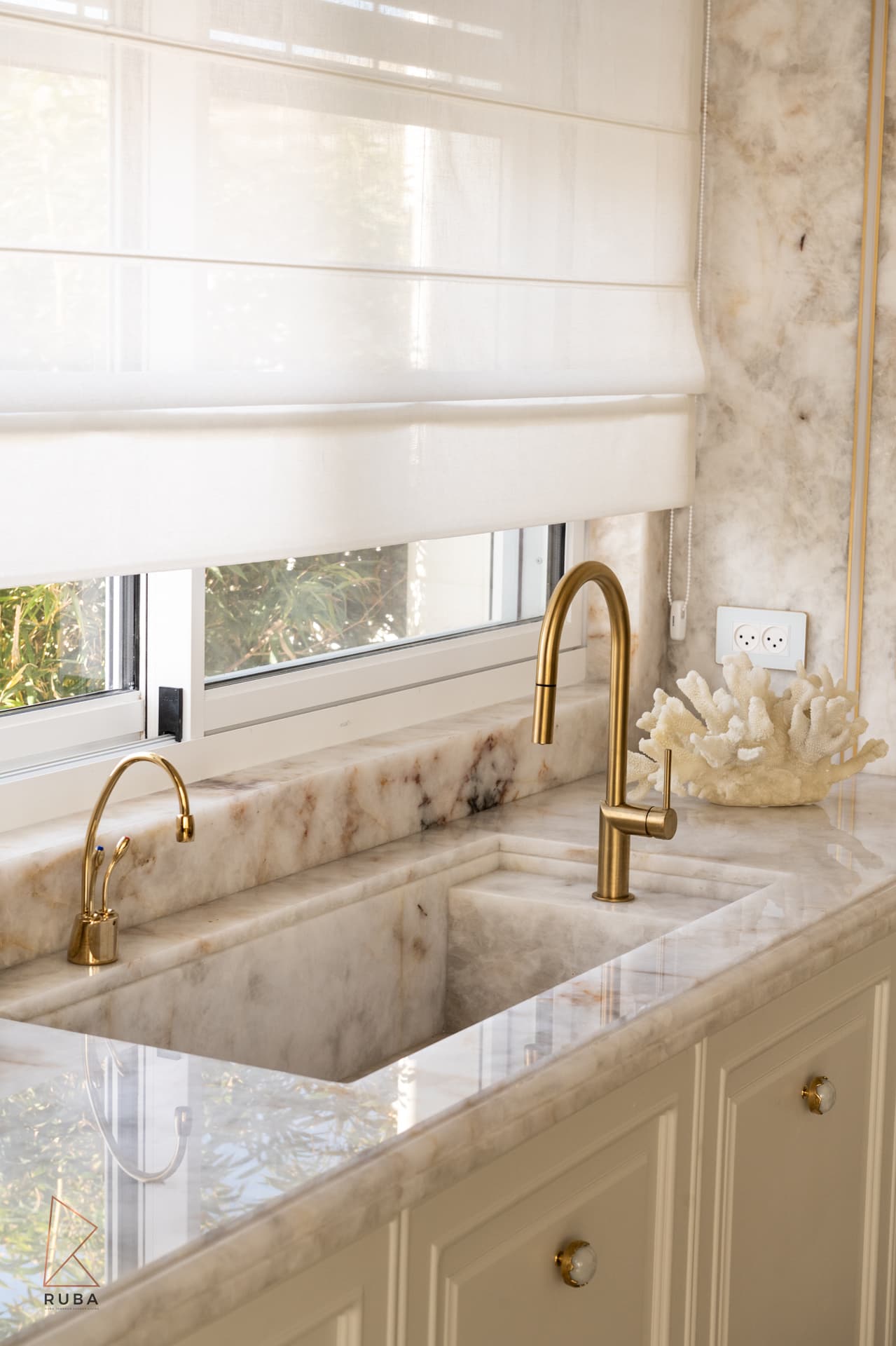 Veined stone sink carved from the same slab as the countertop, with brass faucets beneath a window in the kitchen of The Last Supper