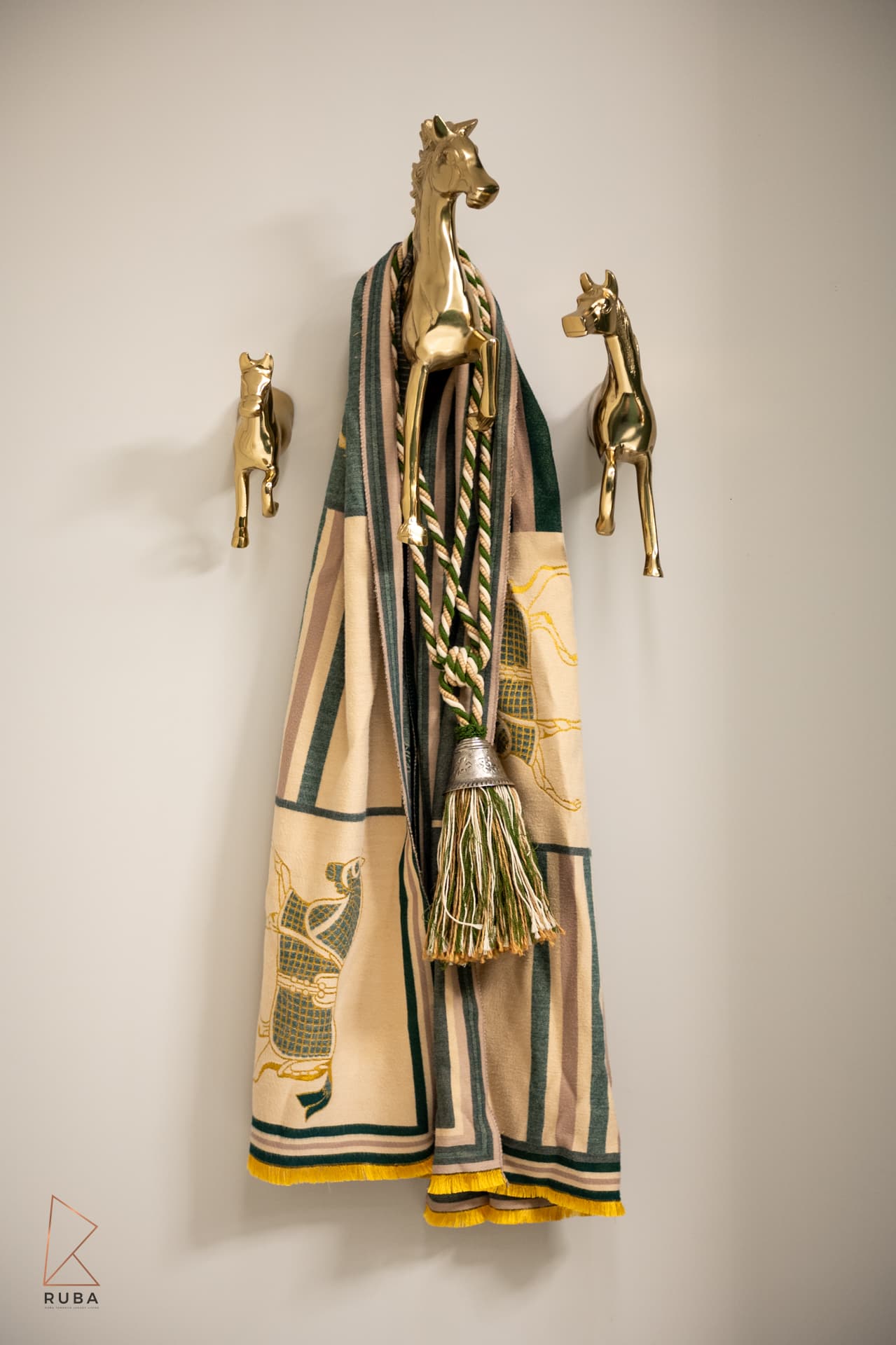 Three brass horse-shaped wall hooks holding a patterned scarf and tasselled cord on a cream wall in The Last Supper