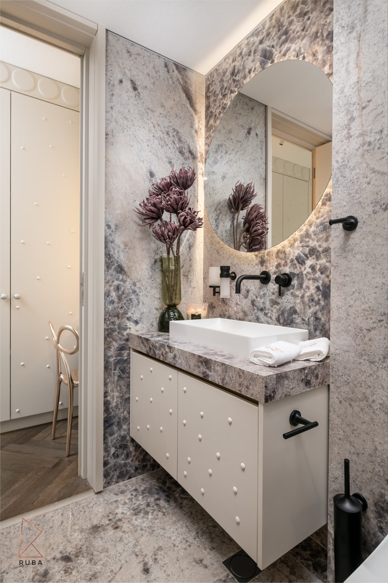Powder room with speckled pink-grey stone walls, backlit oval mirror and studded cream vanity in The Last Supper
