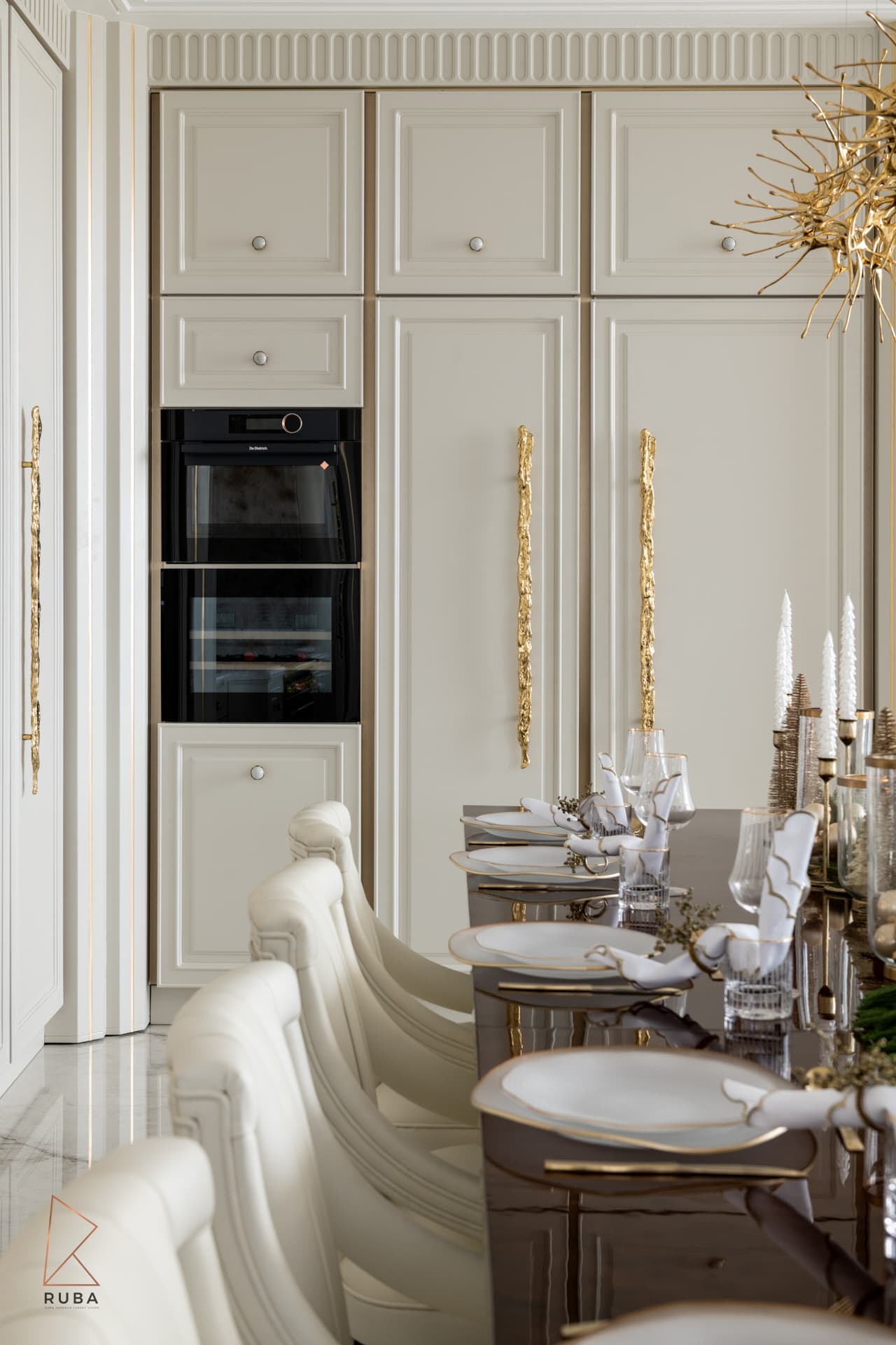 Cream panelled cabinetry with built-in ovens and sculptural brass pulls beside the set dining table in The Last Supper