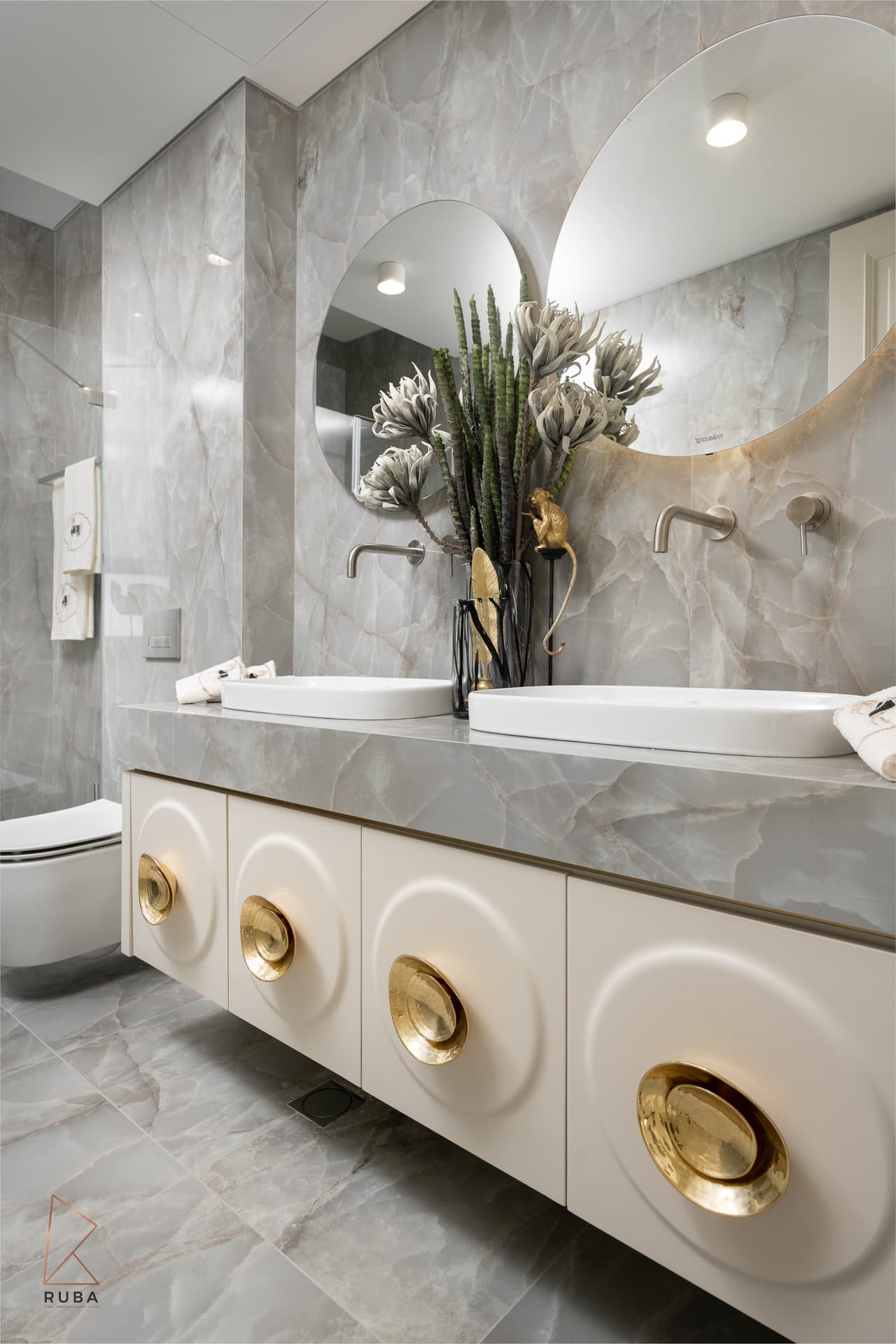 Kids bathroom vanity with circular carved cream doors, brass bowl pulls and a grey veined stone top under round mirrors in The Last Supper