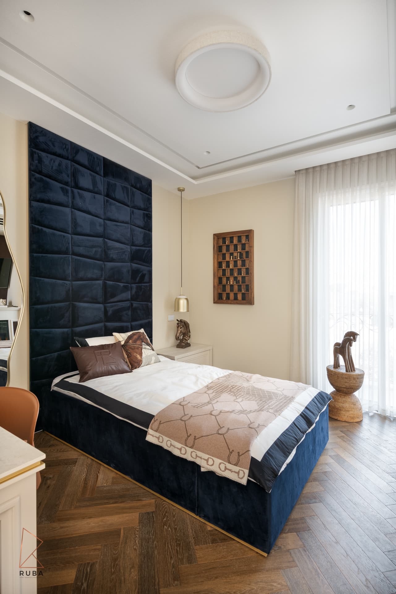 Angled view of a kids bedroom with a navy velvet bed, brass pendant and sheer-curtained window in The Last Supper