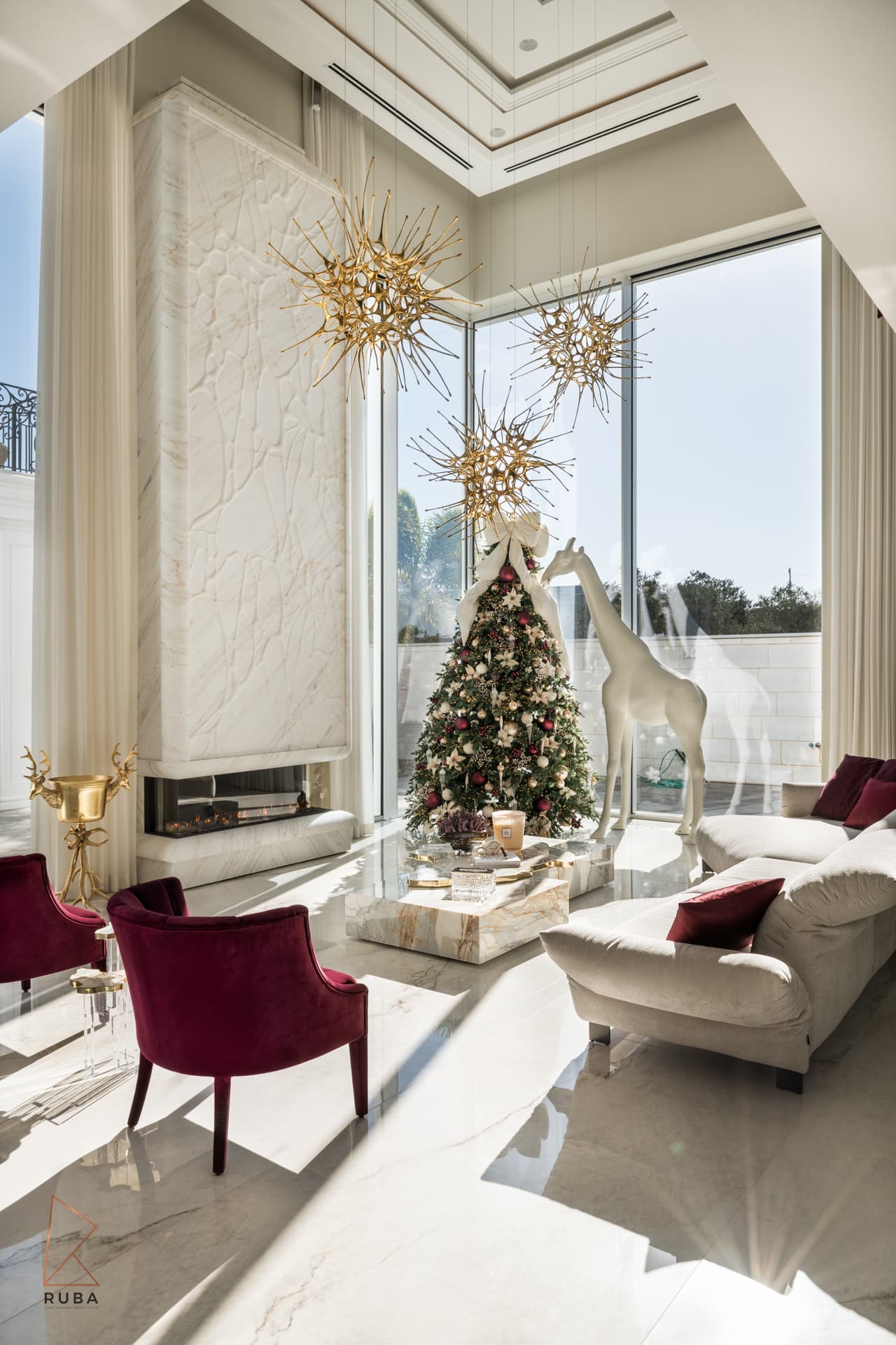 Corner of the double-height living room with tall glazing, a veined stone fireplace column and burgundy velvet armchairs in The Last Supper